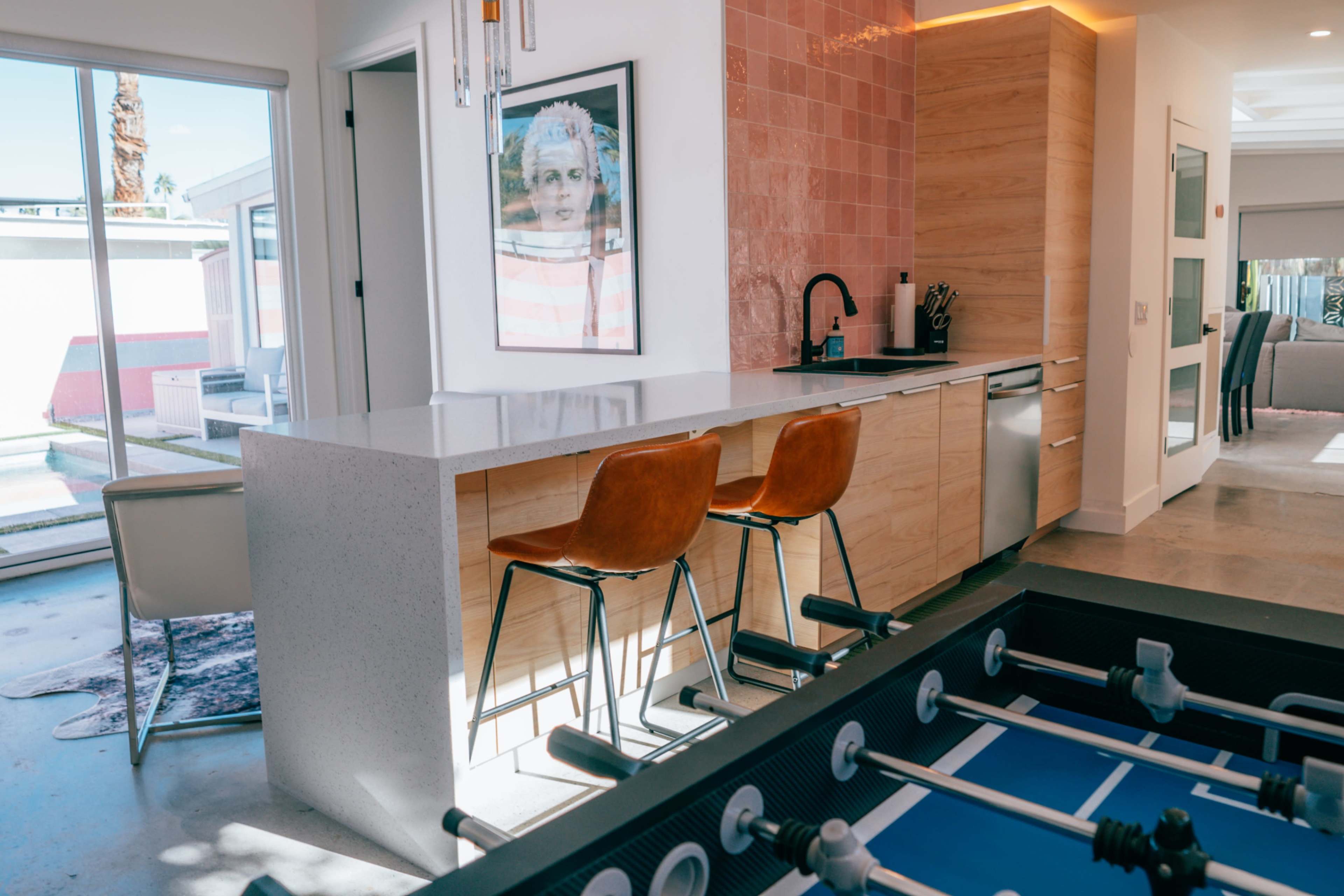The image shows a modern kitchen with a countertop, wooden cabinets, and two brown bar stools, adjacent to a foosball table and a view of an outdoor pool area.
