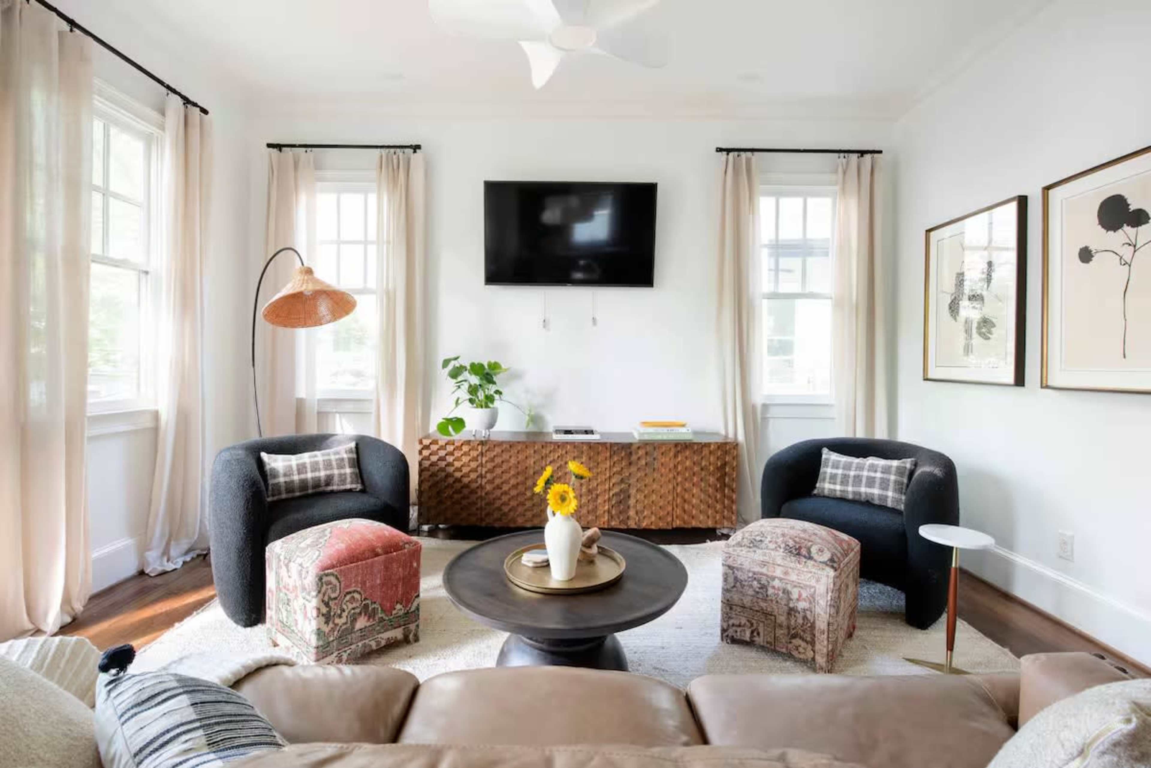 The image shows a modern living room with two armchairs, a round coffee table, a television mounted on the wall, and decorative elements including a plant and framed artwork.