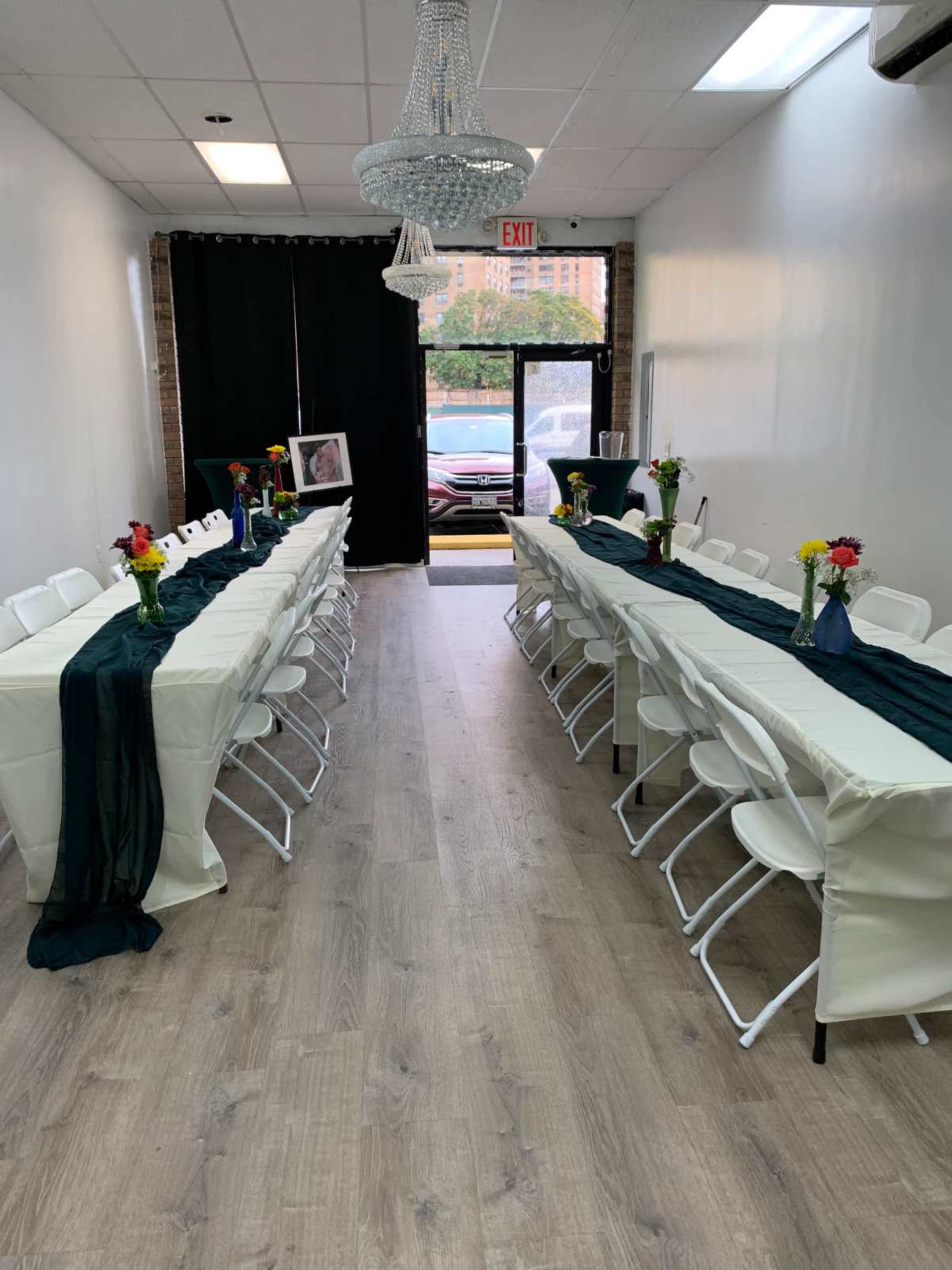 The image shows a long, narrow room with two rows of tables set for an event, each adorned with a dark table runner and a vase of flowers.