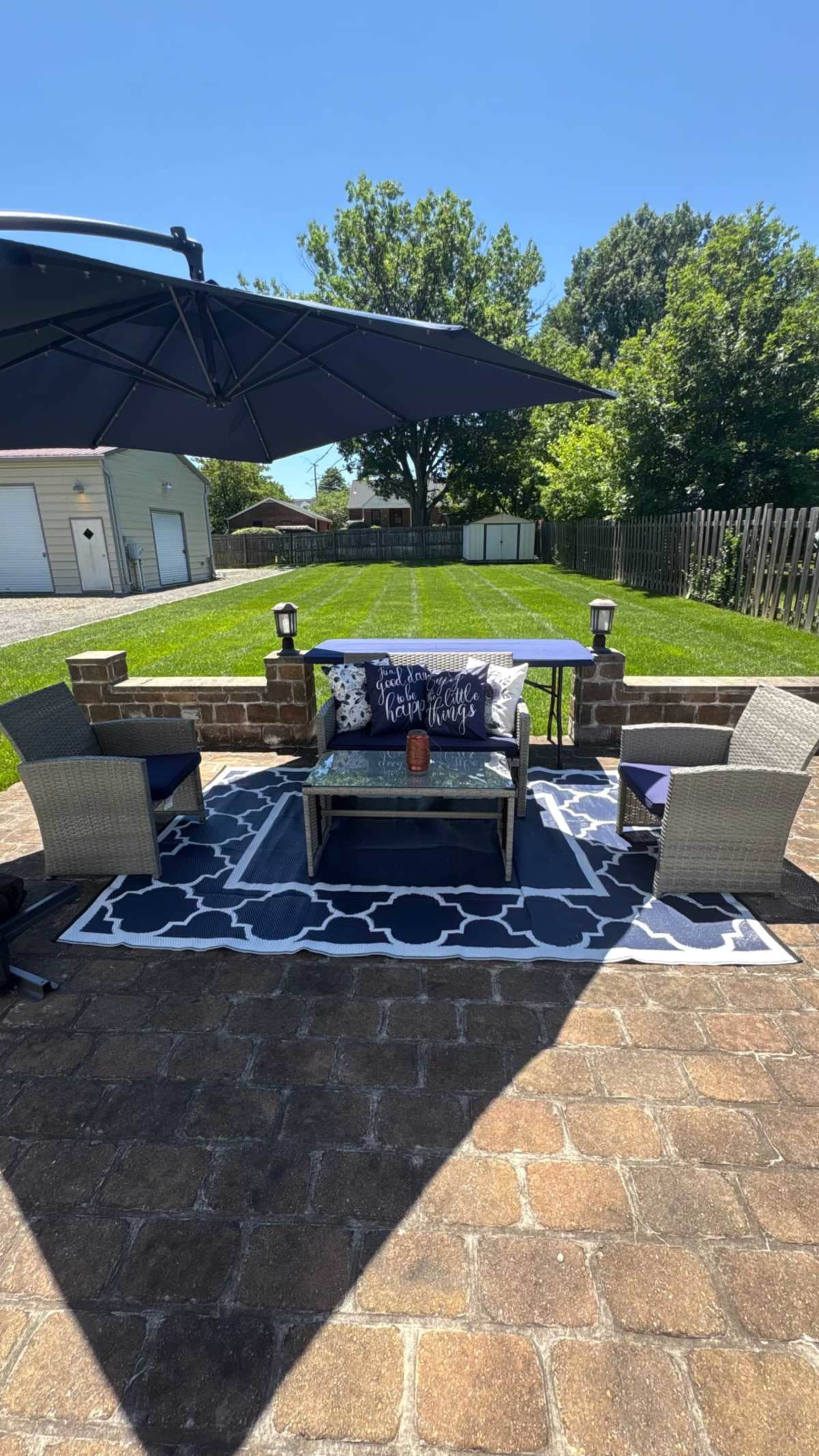 The image shows a patio area with two chairs, a coffee table, and a large umbrella, surrounded by a neatly maintained lawn and a wooden fence.