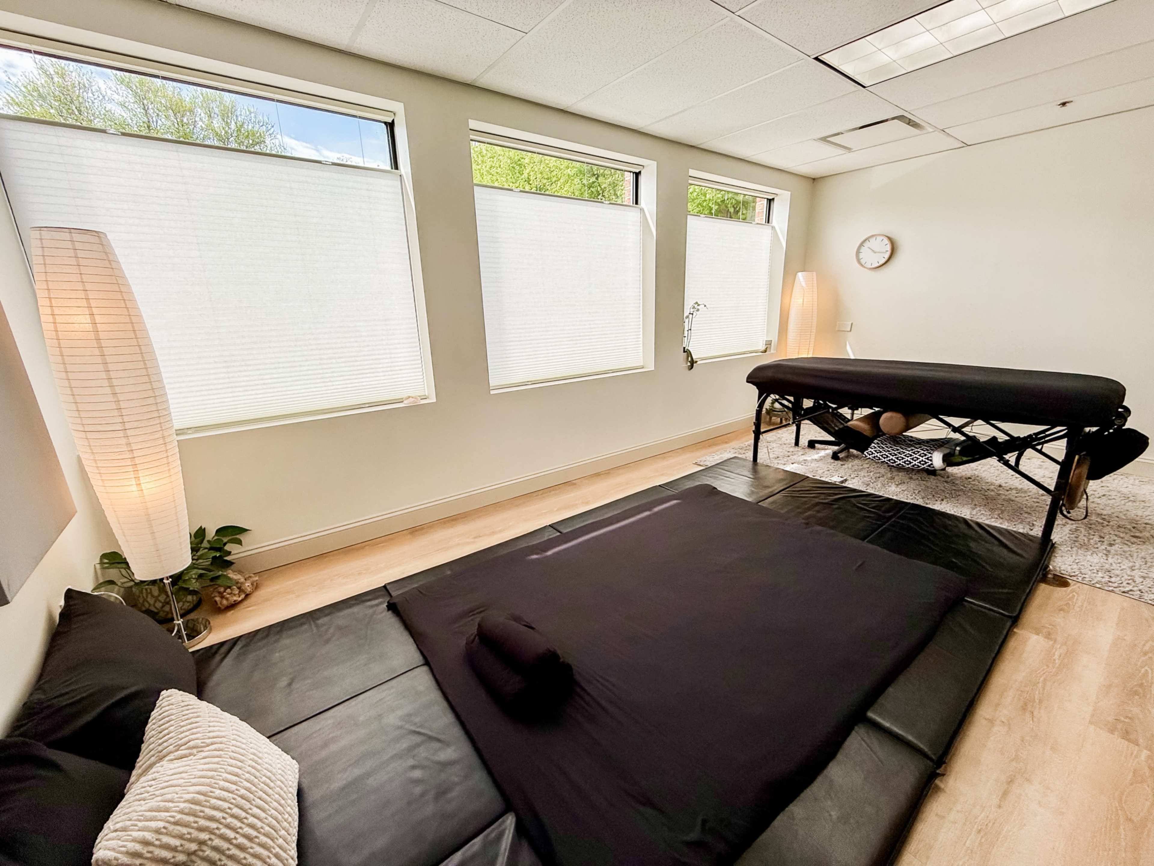 The image shows a spacious therapy room with large windows, a massage table, floor cushions, and soft lighting.