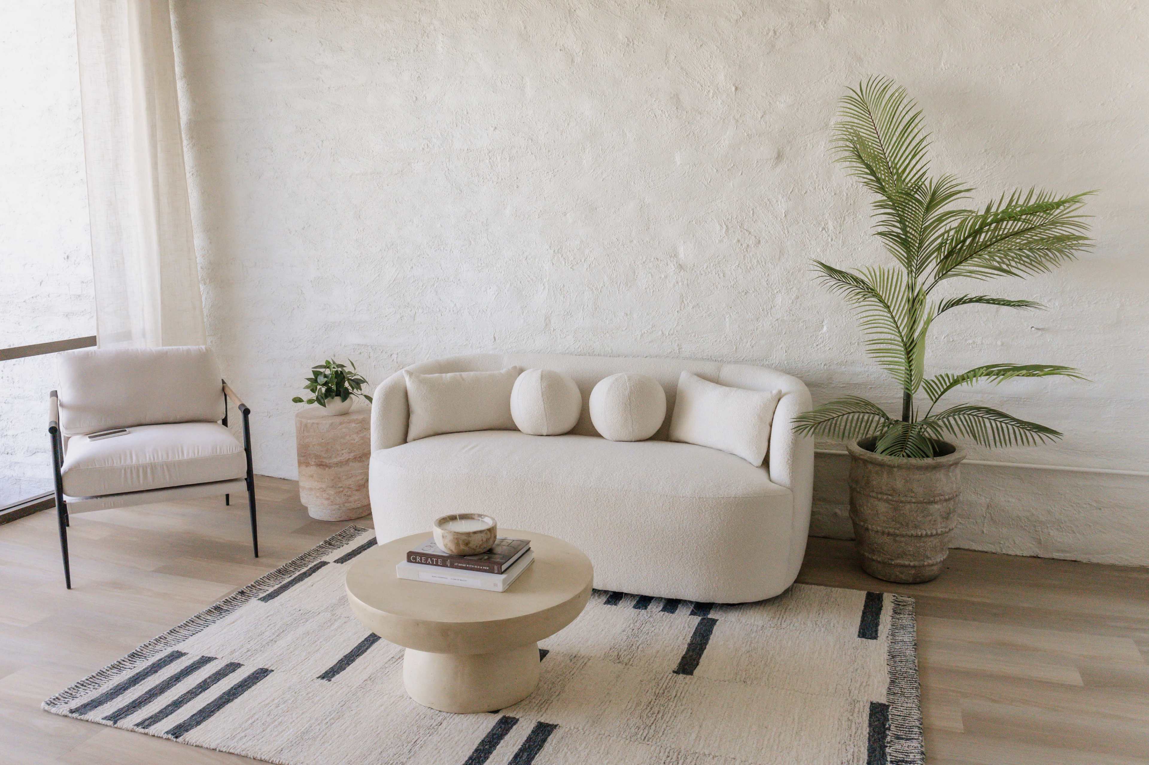 The image shows a modern living area featuring a rounded white sofa with three circular cushions, a minimalist coffee table, an armchair, and a large potted plant against a textured white wall.