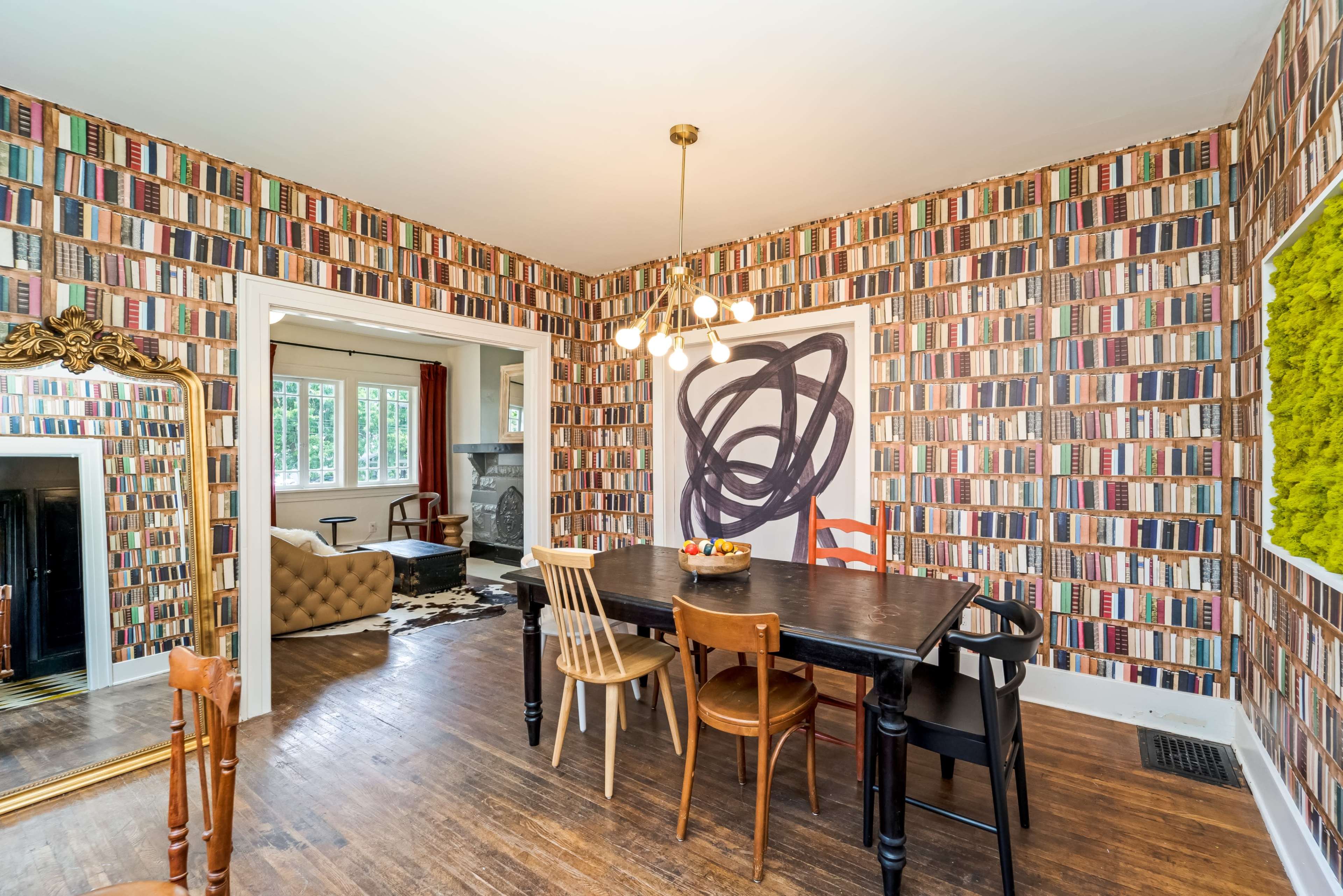 A dining area features a dark table surrounded by chairs, with a wall adorned with book-patterned wallpaper and an abstract art piece.
