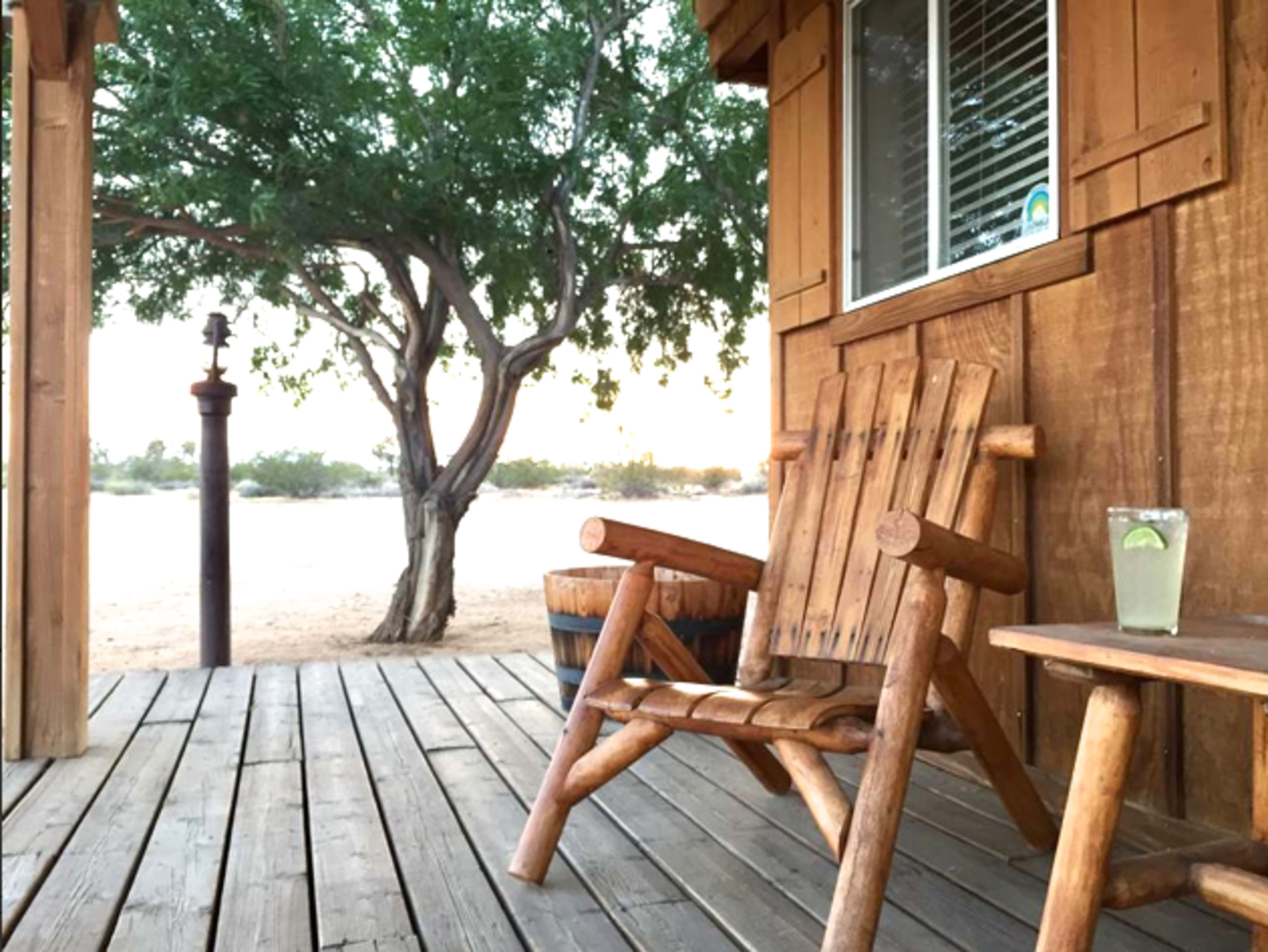 A wooden deck features two rustic chairs, a side table with a drink, and a tree nearby, set against a sandy backdrop.