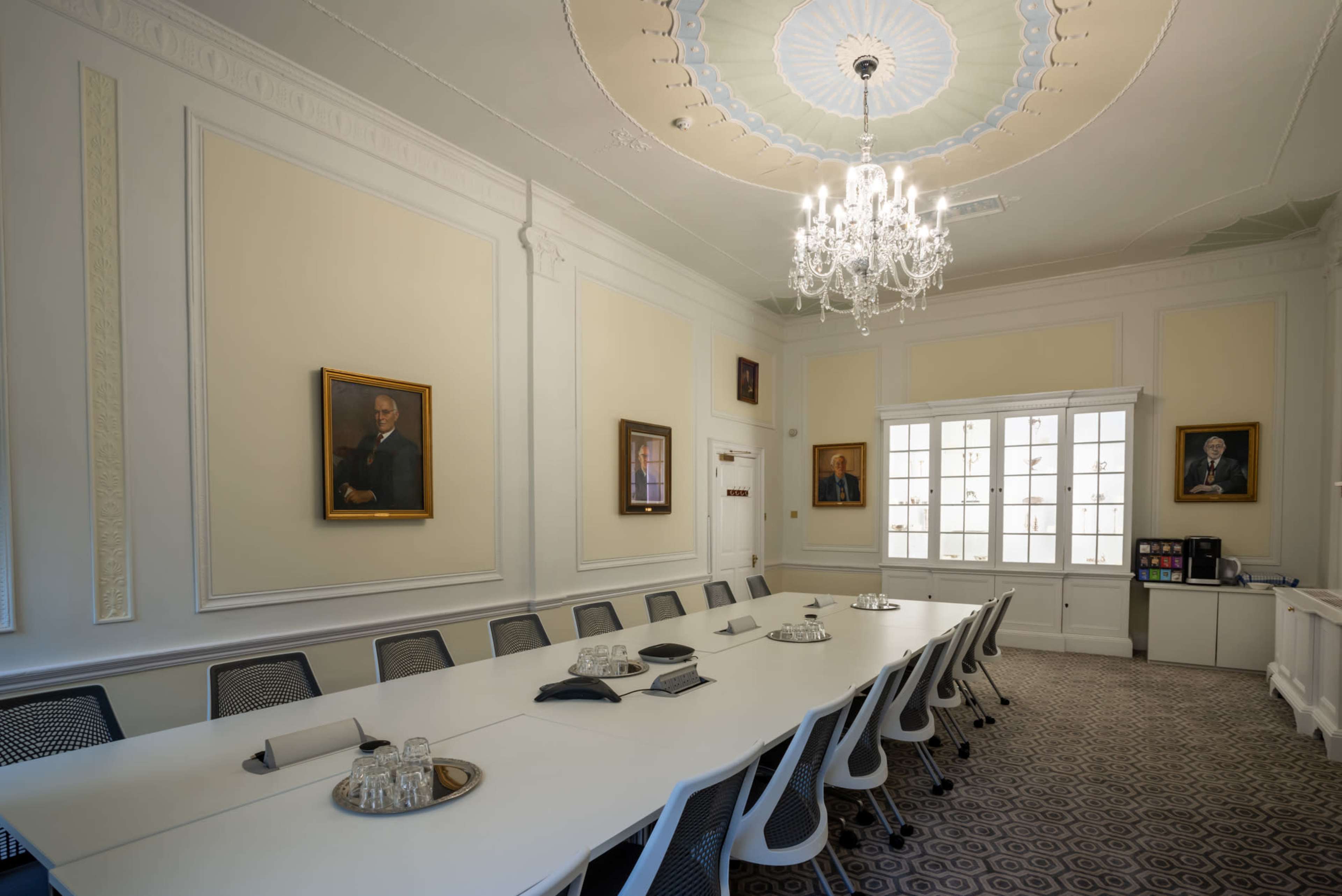 A long, modern conference table is set in a well-lit meeting room adorned with framed portraits and a chandelier.
