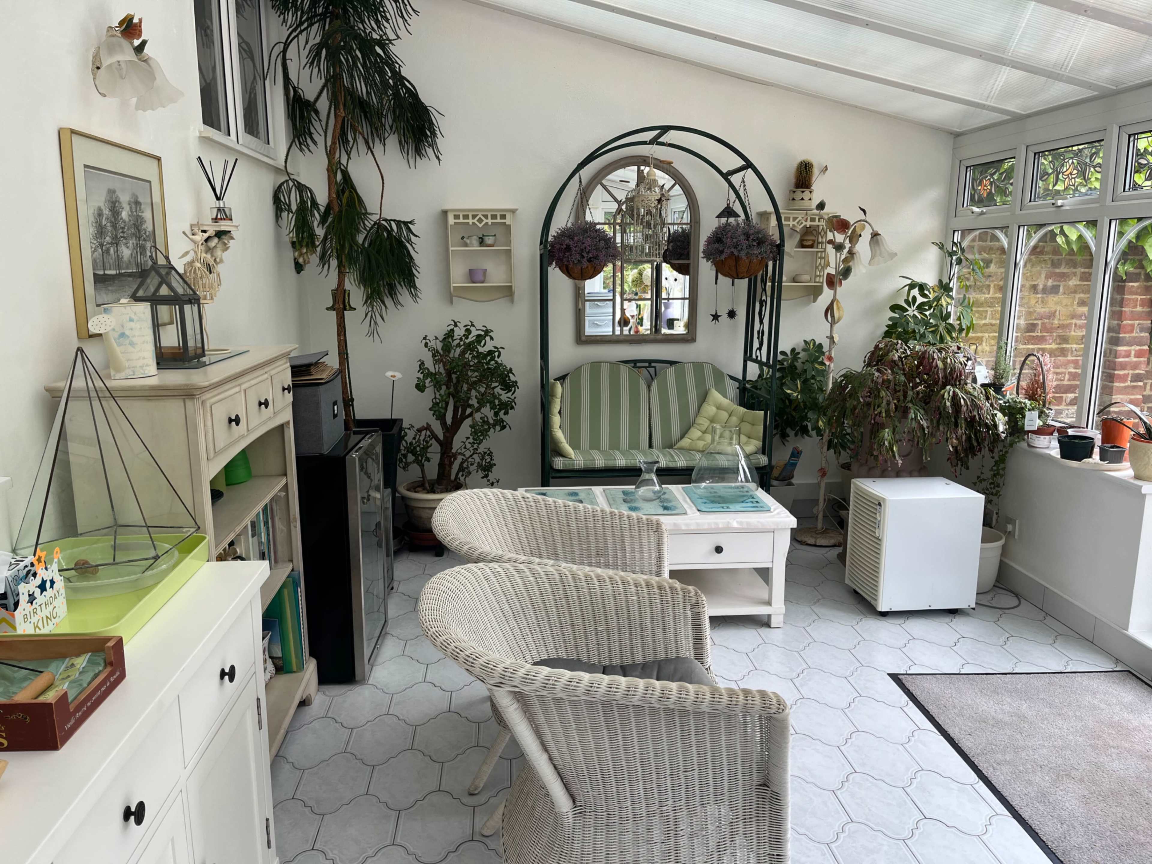 The image depicts a sunlit room with plants, a seating area, and various decorative elements, creating a cozy indoor space.