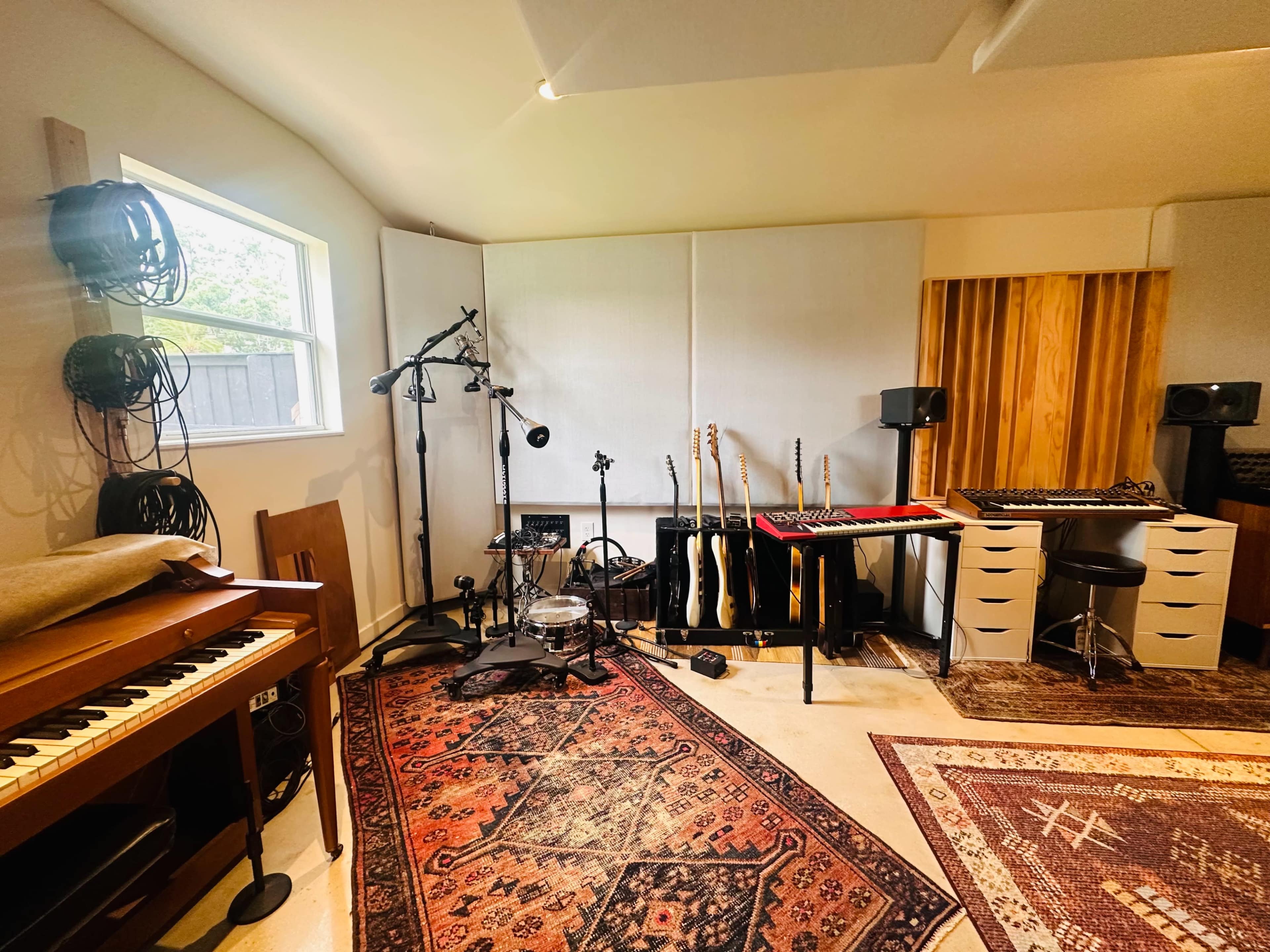 The image shows a music studio equipped with various musical instruments, including guitars, a keyboard, and recording equipment, set against a backdrop of rugs and soundproofing panels.