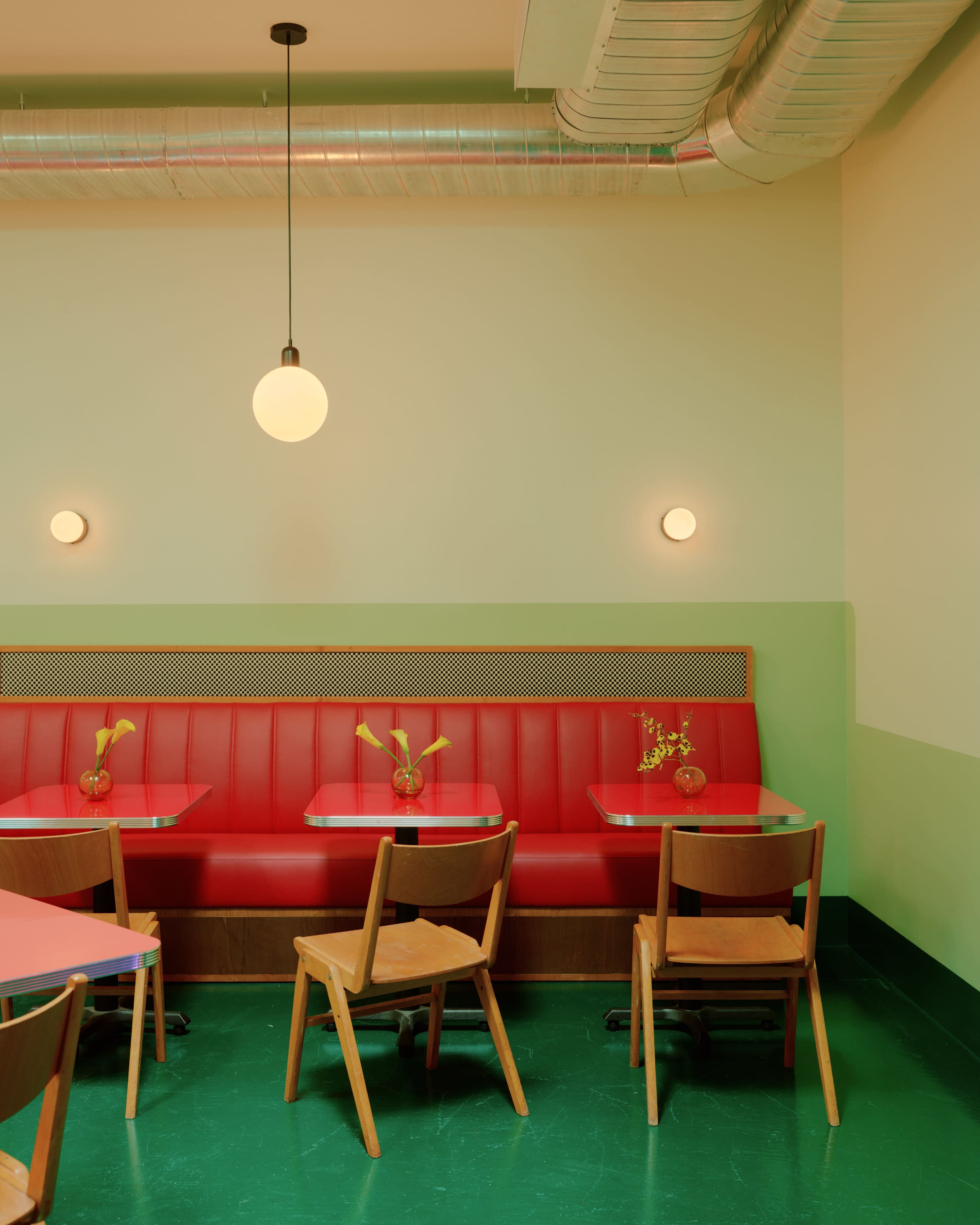 A modern café interior featuring red booth seating, small tables with vases of flowers, and wooden chairs against a green floor and walls.