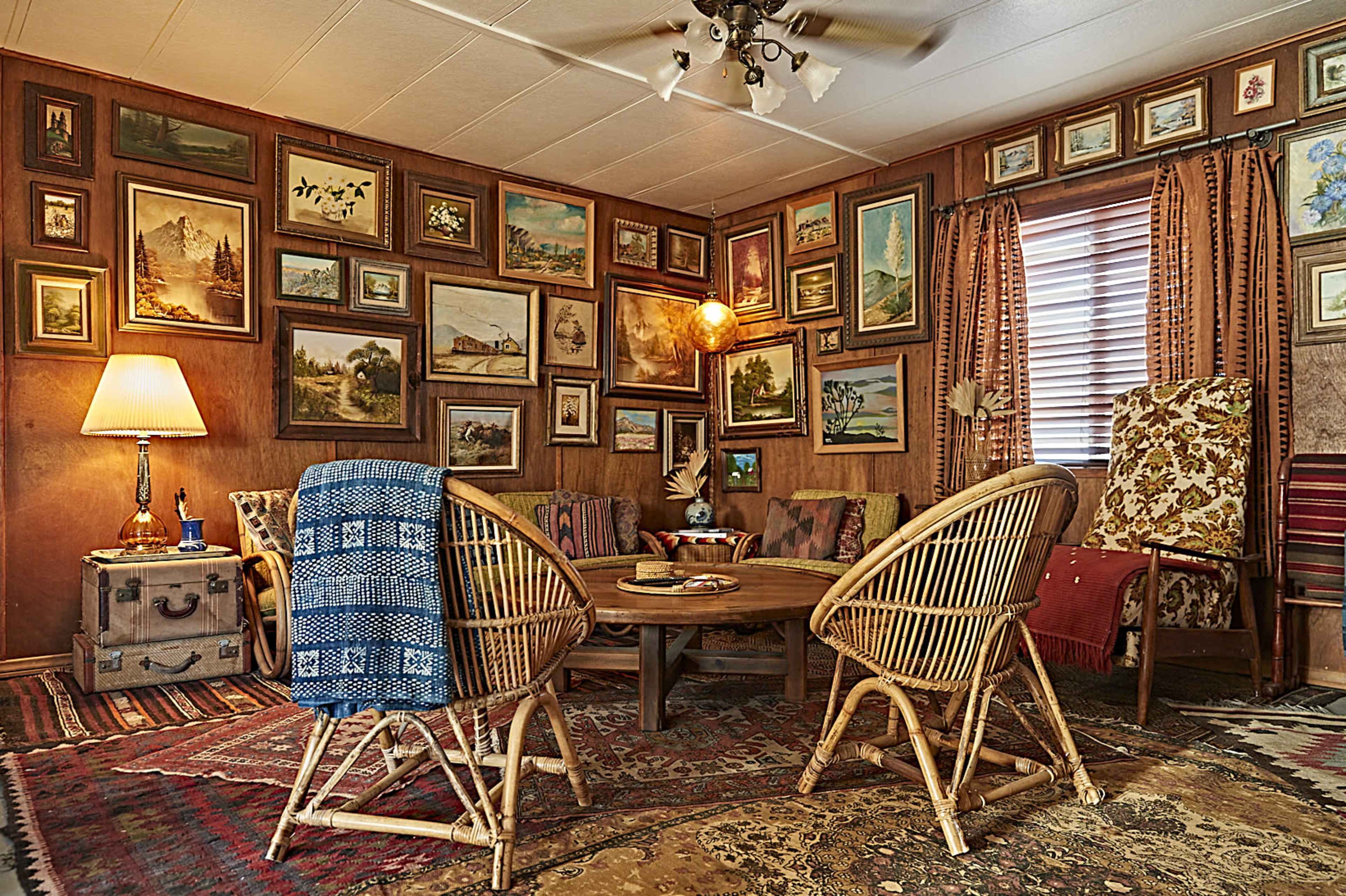 A cozy living room with a round table surrounded by rattan chairs, walls adorned with a variety of framed paintings, and warm lighting coming from a lamp.