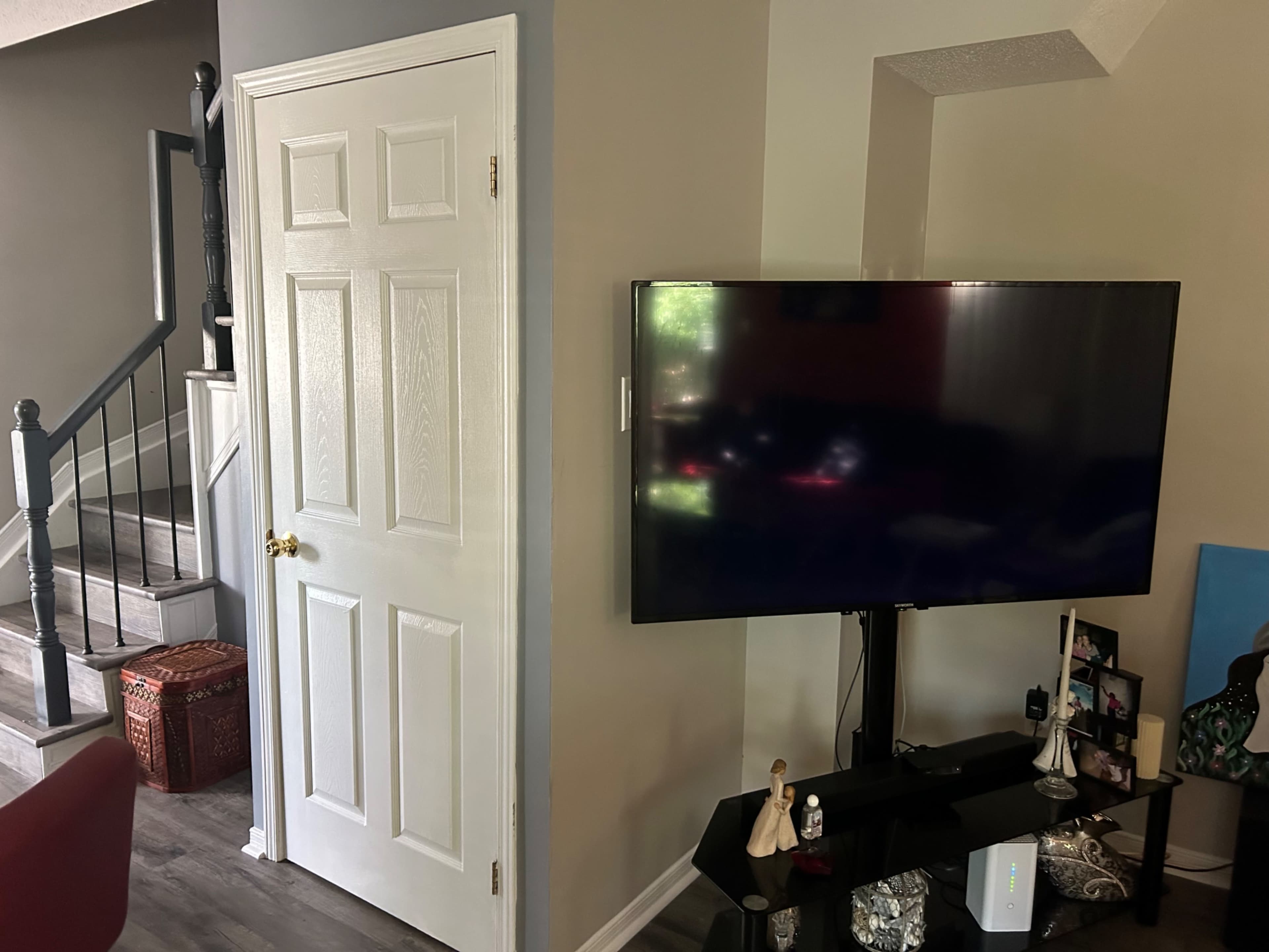 A living room corner with a white door, a large television mounted on a black stand, and a small decorative shelf with items underneath the TV.