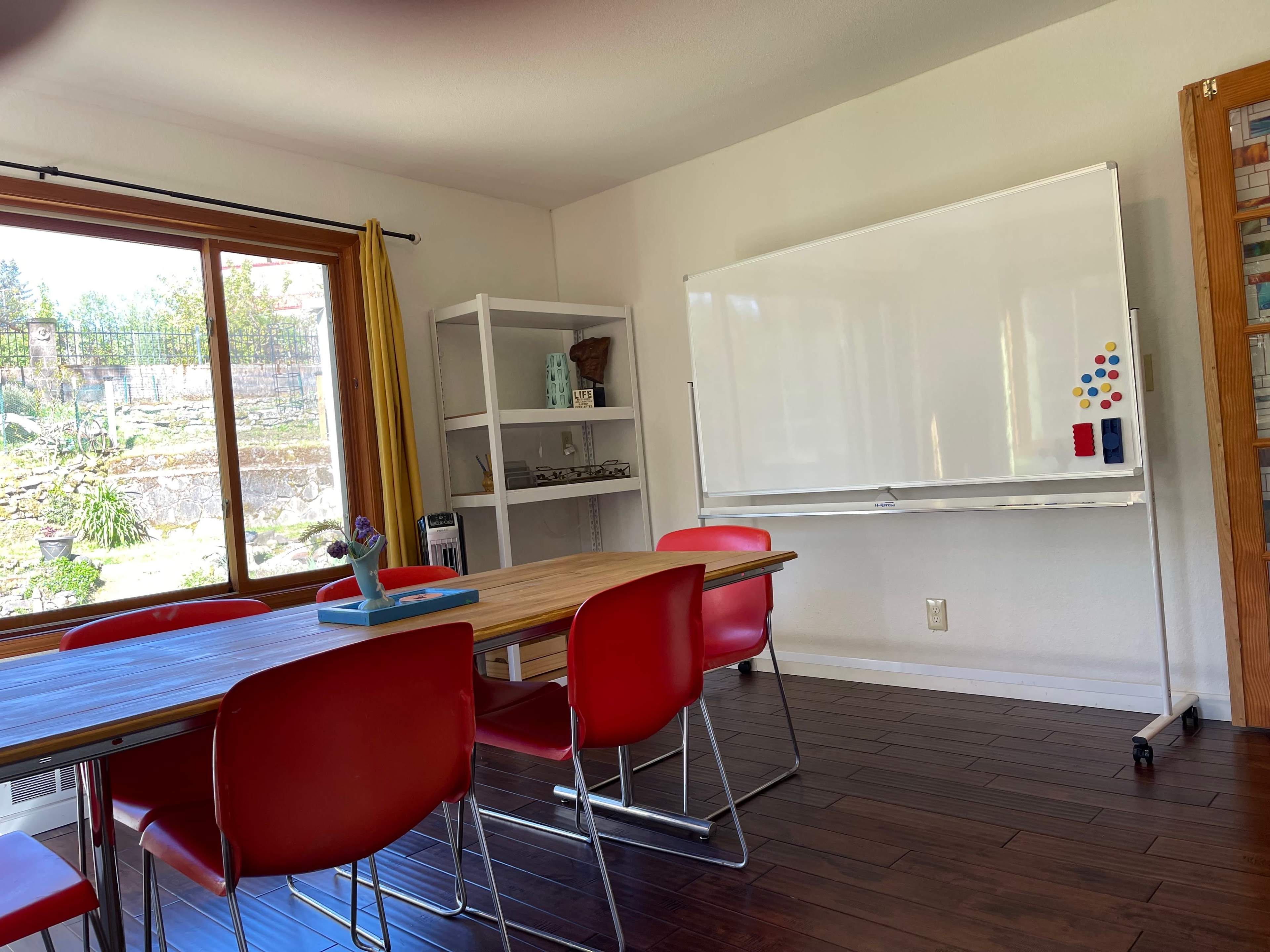 A modern meeting room features a wooden table surrounded by red chairs, with a whiteboard mounted on the wall and a large window overlooking a landscaped area.