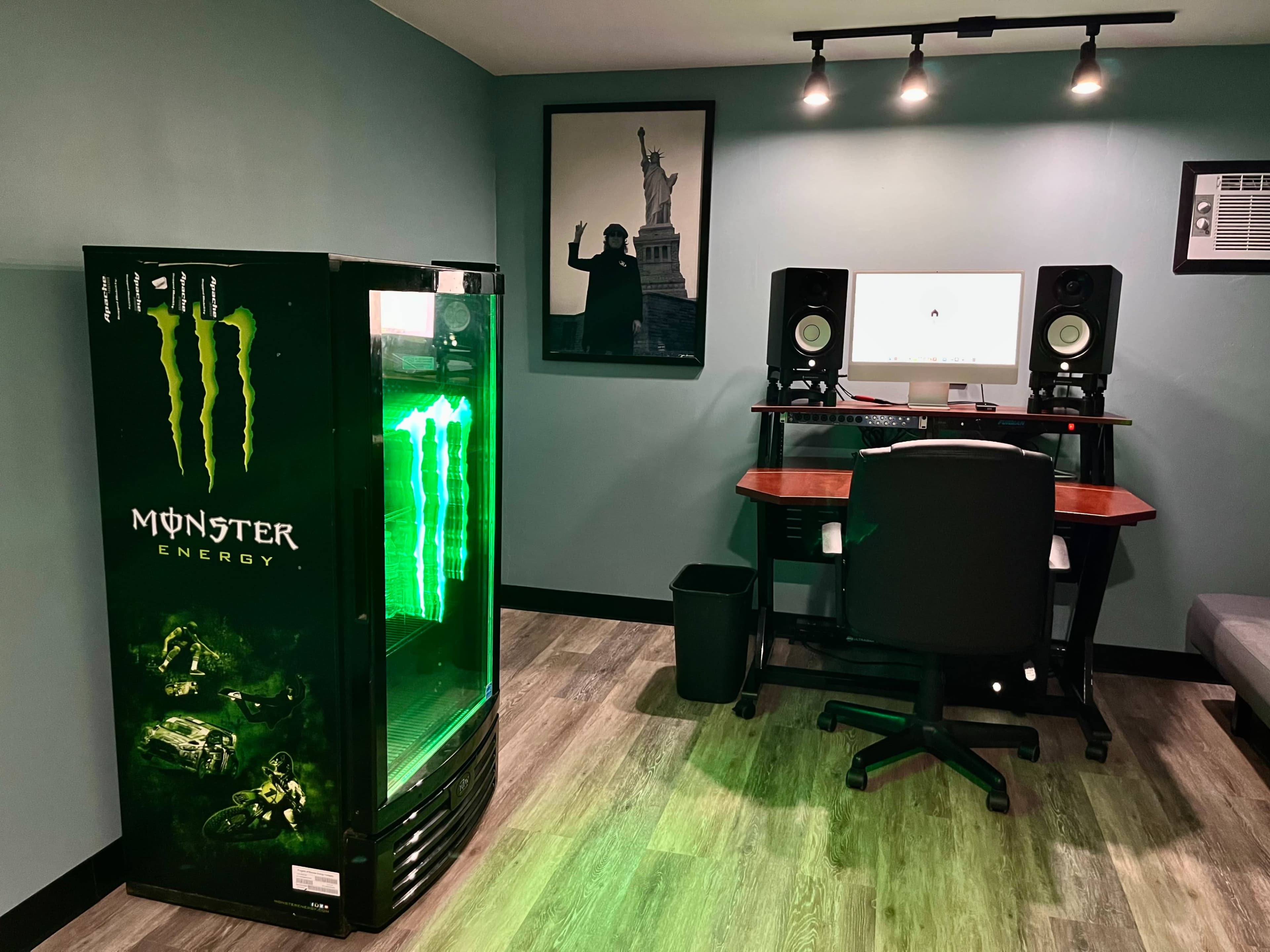 A room with a Monster Energy drink fridge on the left, a desk with a computer and speakers in the center, and a wall featuring a framed picture of the Statue of Liberty.