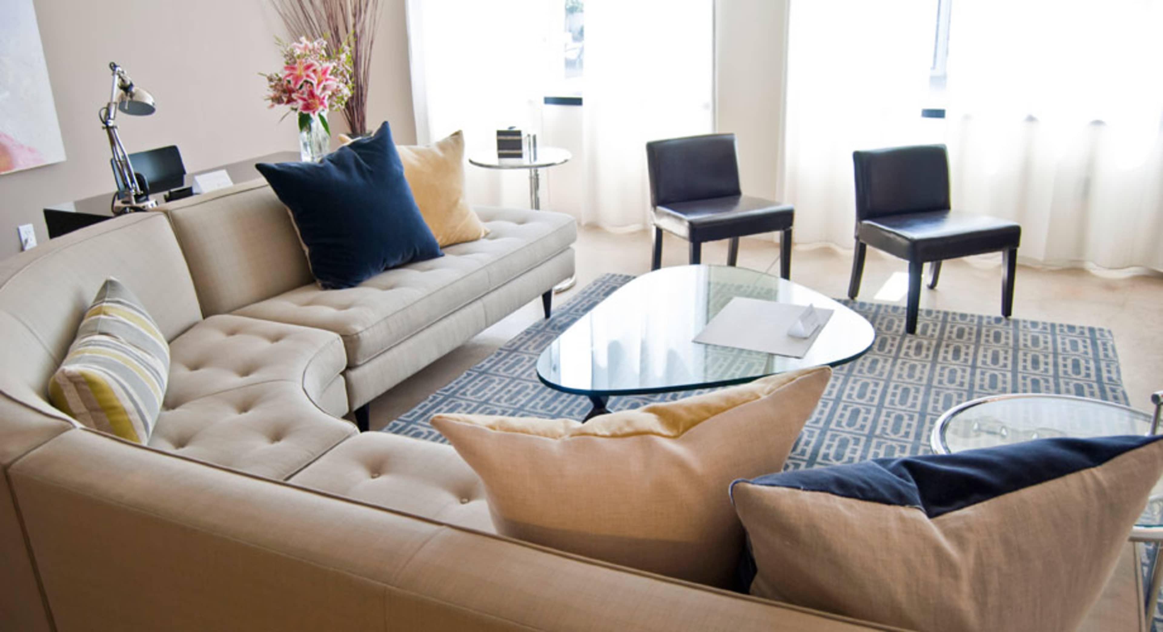 A modern living room with a sectional sofa, a glass coffee table, and a patterned rug, accompanied by chairs and large windows allowing natural light.