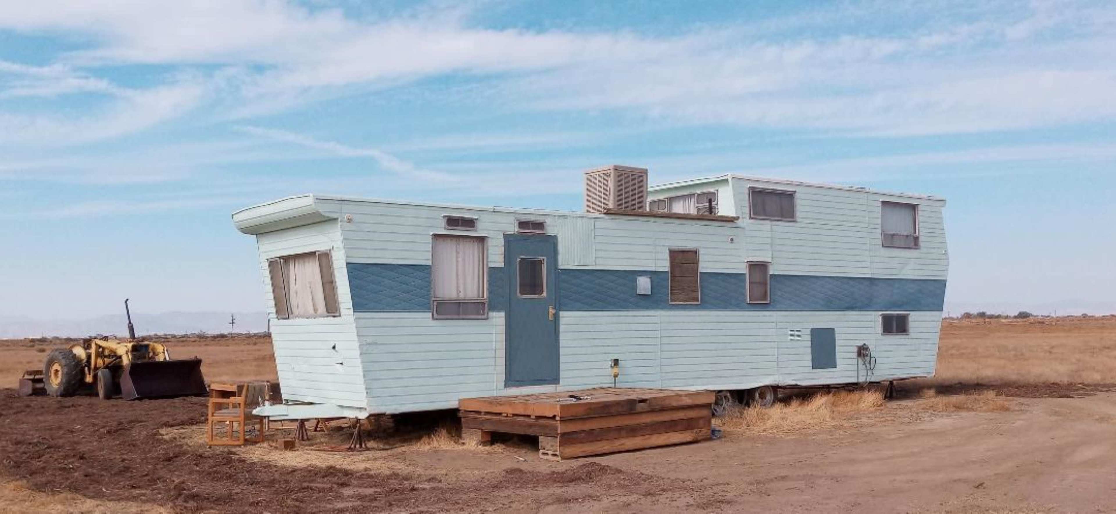 A light blue mobile home is parked on a dirt patch in an open field with a yellow tractor nearby.