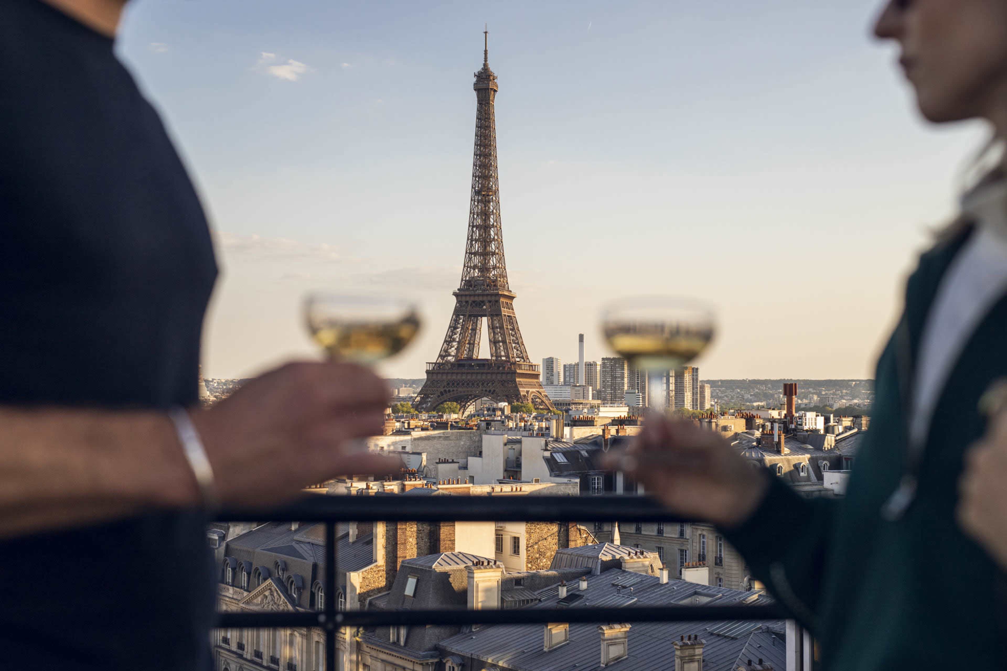 Two people hold champagne glasses while overlooking a view of the Eiffel Tower in Paris at sunset.