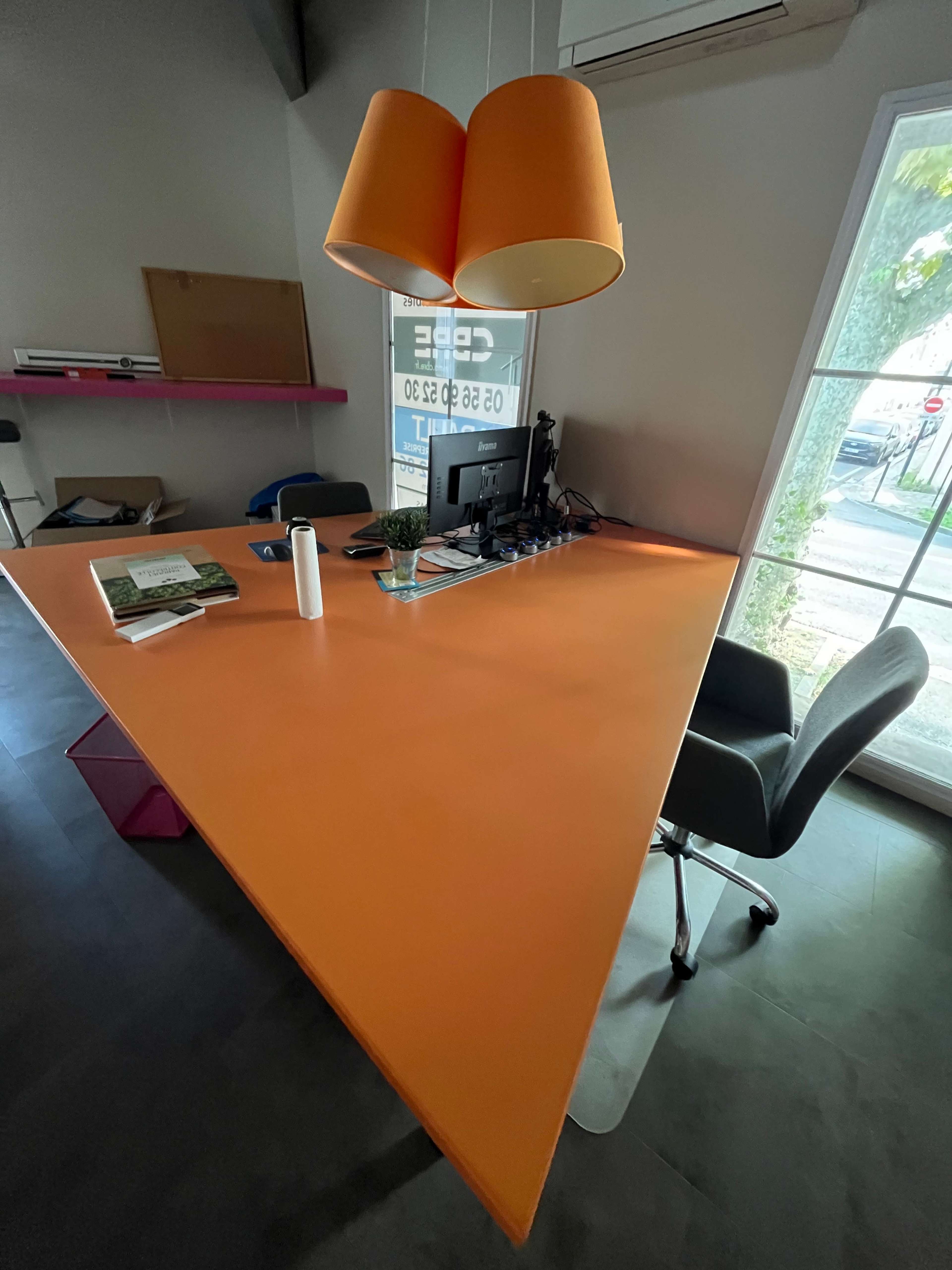 The image shows a modern office space with a triangular orange desk, a chair, a computer setup, and large windows providing natural light.