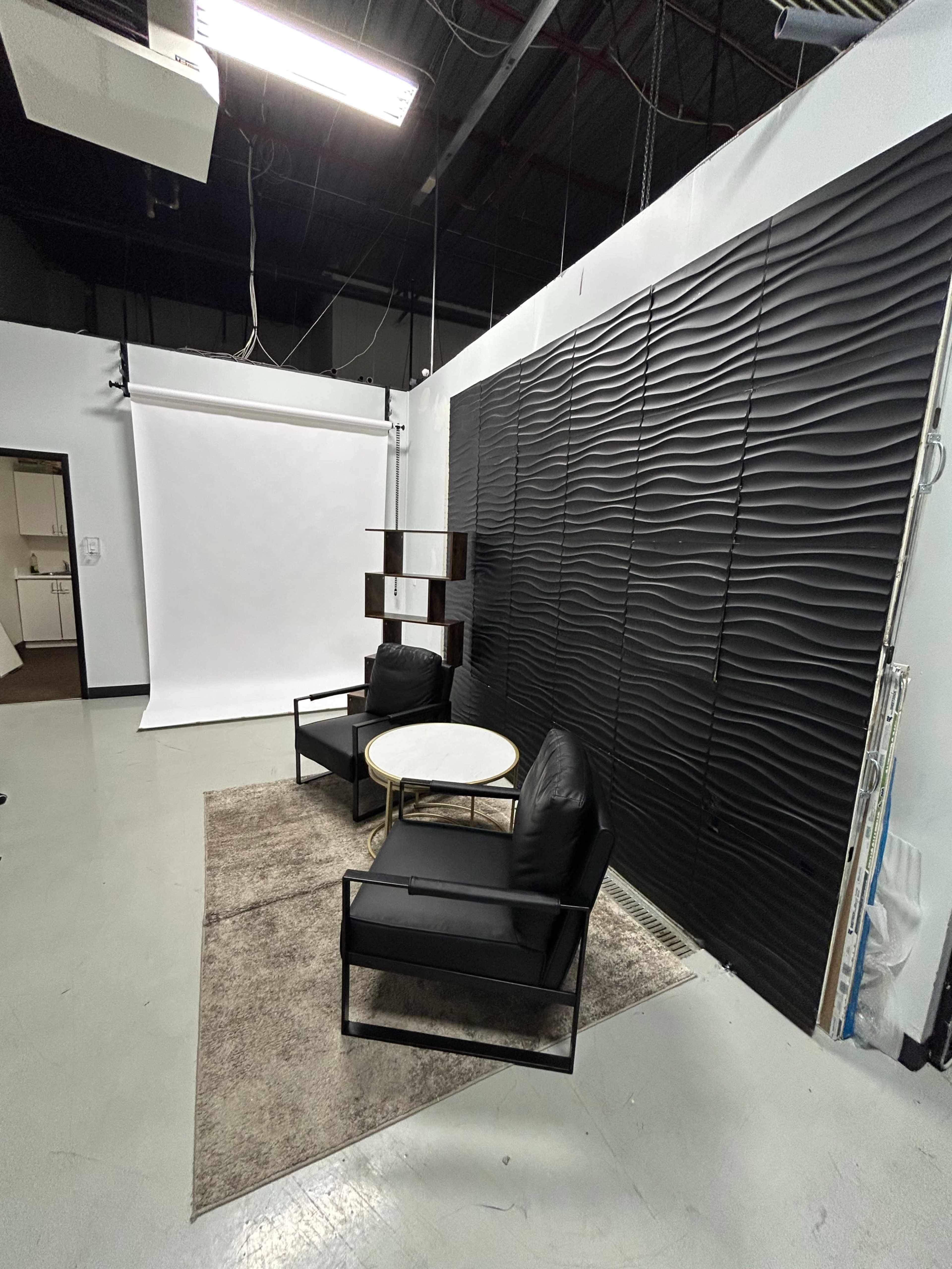 The image shows a minimalist studio interior featuring two black chairs and a round table on a rug, with a textured black wall and a white backdrop in the background.