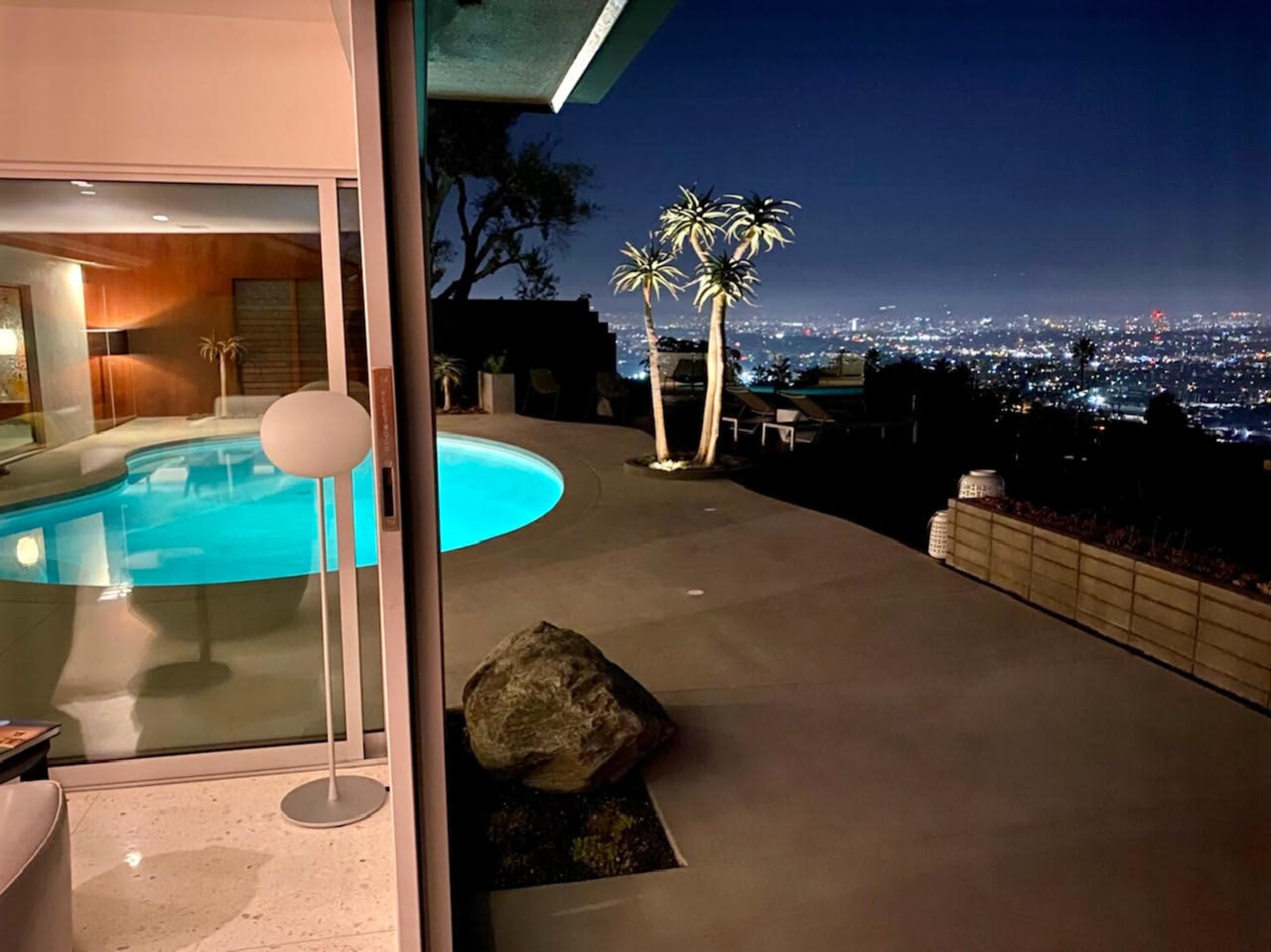 A swimming pool glows blue under the night sky, with city lights visible in the distance and a modern home partially illuminated in the foreground.