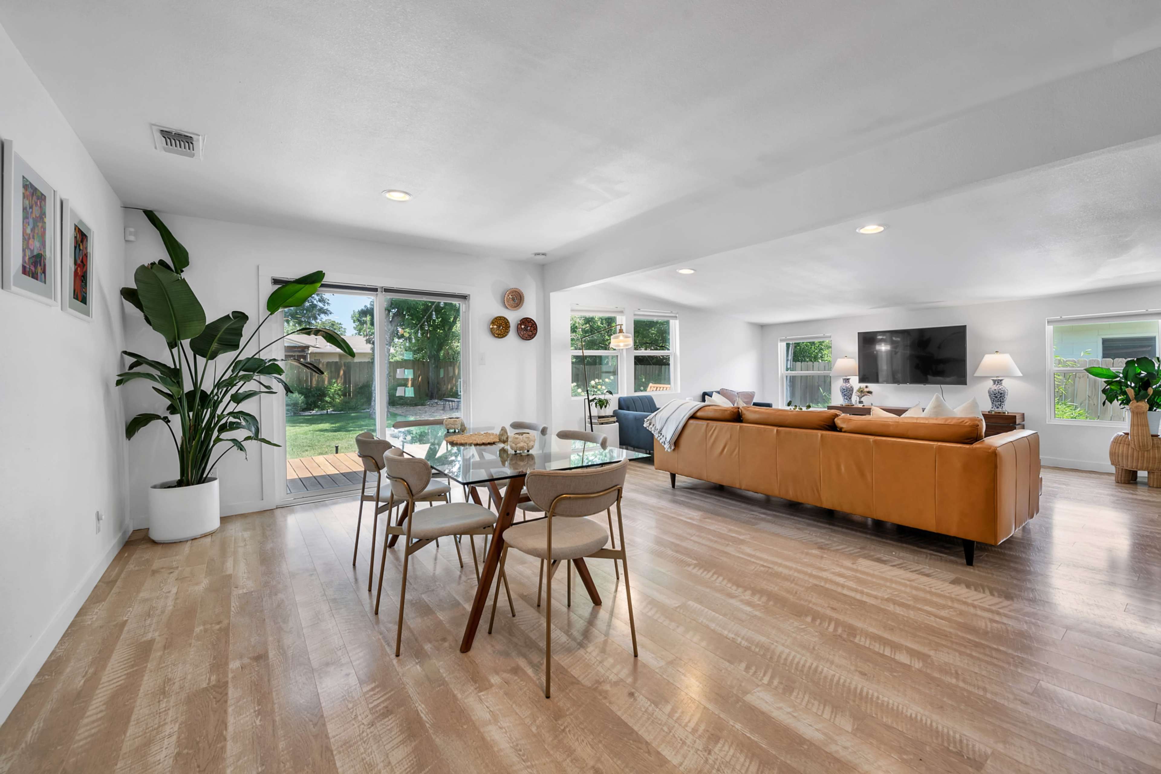 The image shows a modern living space with a dining area featuring a glass table and four chairs, a brown leather sofa, and large windows that provide natural light.