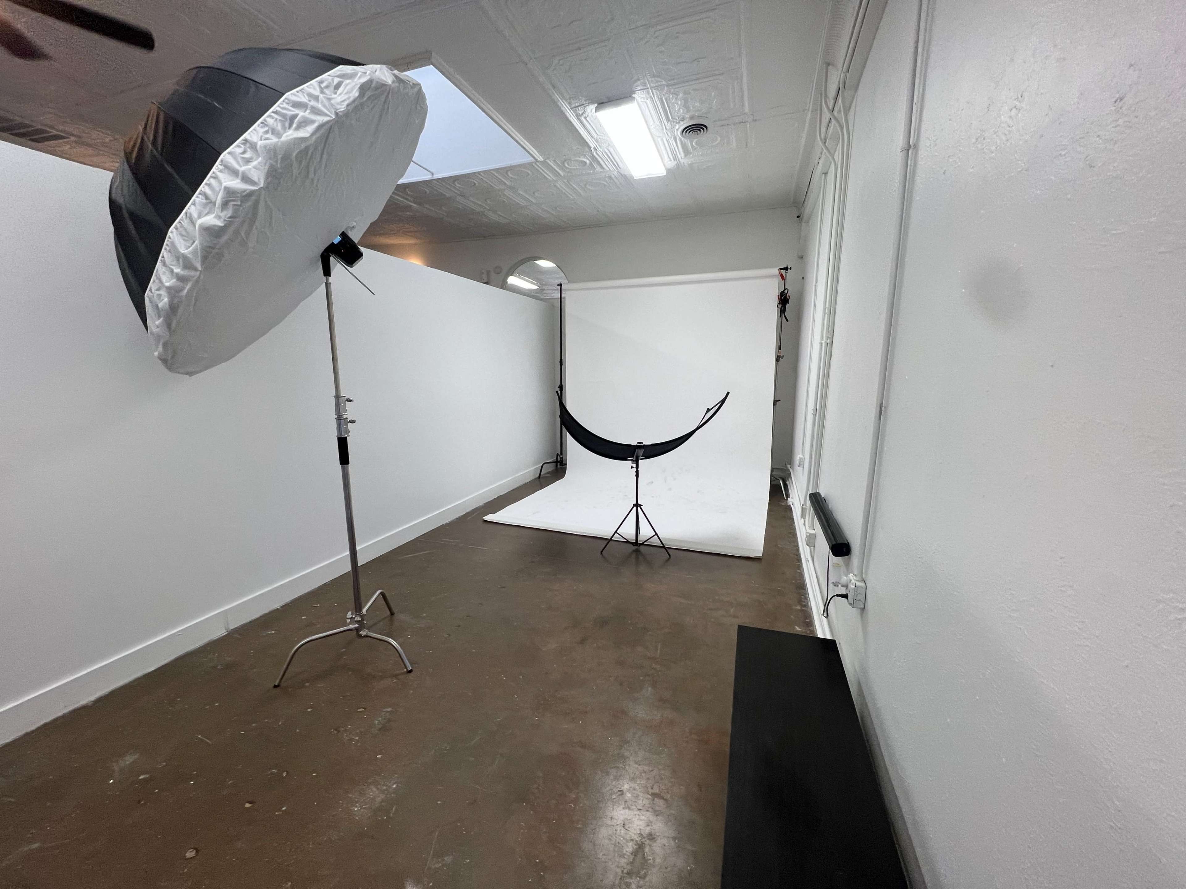 The image shows a photography studio with a large umbrella softbox, a backdrop stand, and a plain white backdrop set up on a concrete floor.
