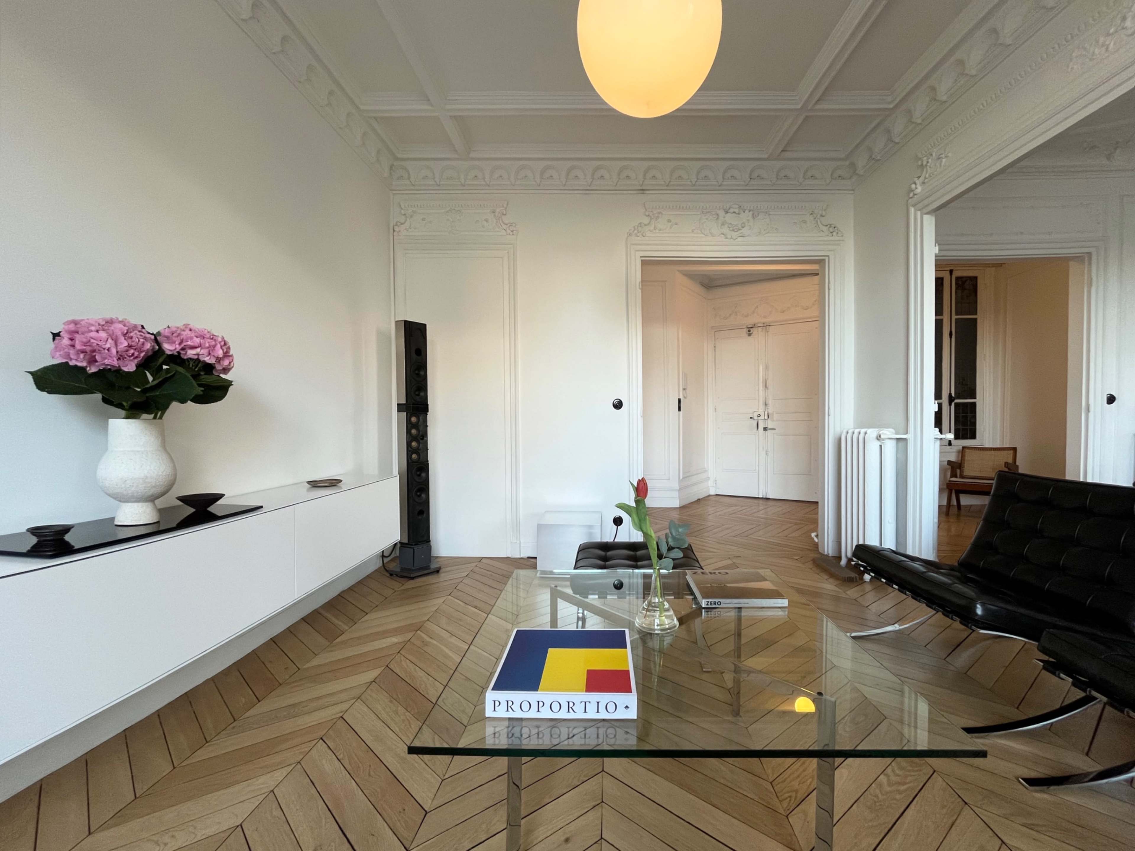 A modern living room features a glass coffee table, a black leather chair, and decorative elements like flowers and a sign, all set against a backdrop of elegant white walls and a hardwood floor.