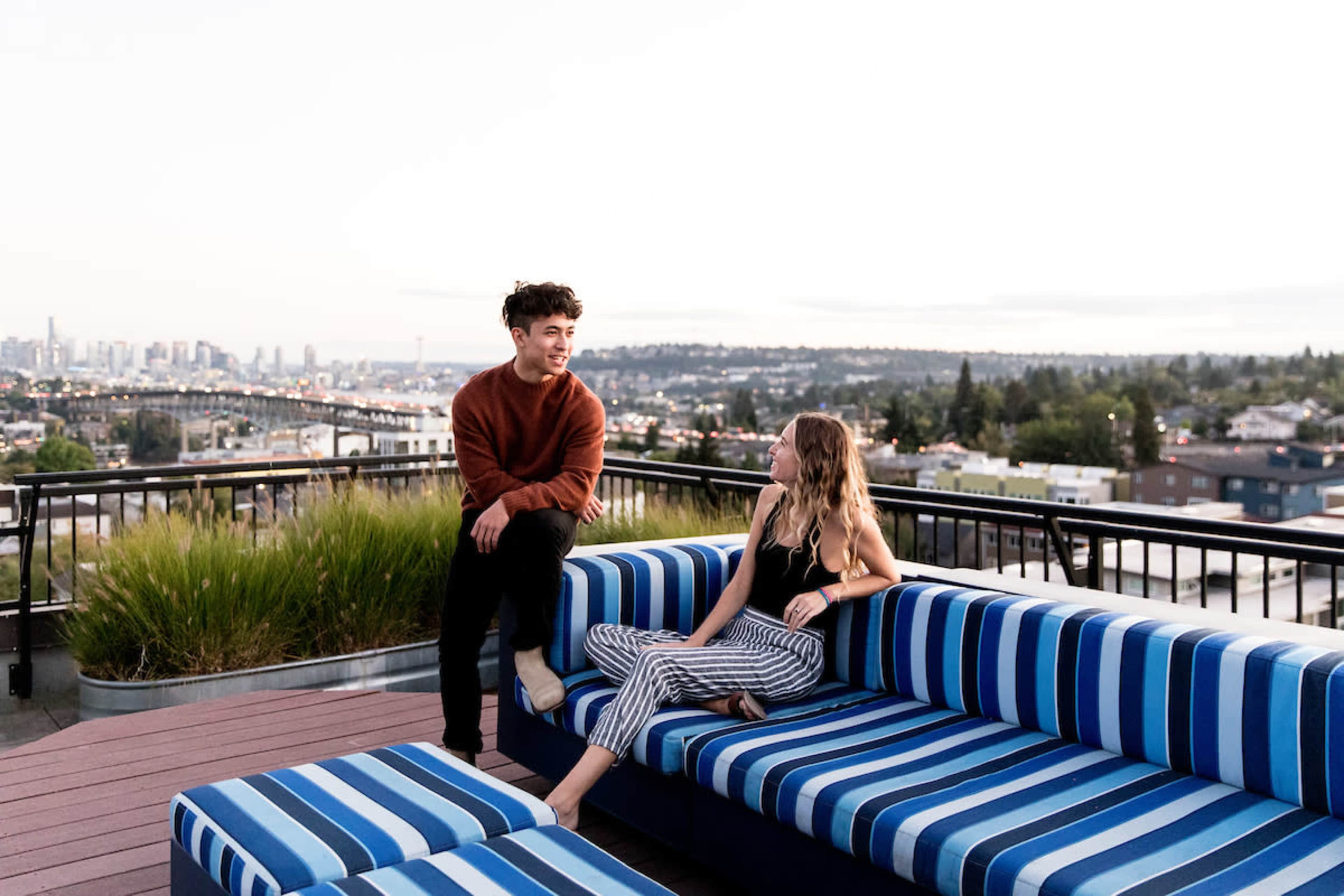 A young man in a sweater sits on a striped couch while a young woman in patterned pants leans back, both looking at each other with a city skyline in the background.