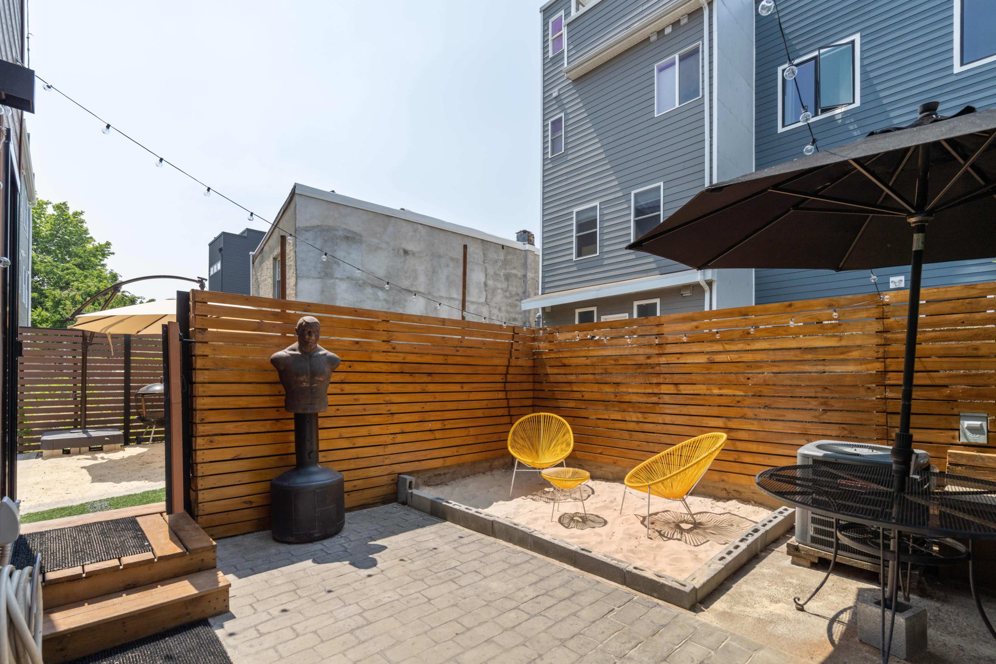 The image shows a small outdoor patio area featuring a sand pit, two yellow chairs, a black umbrella, and a wooden privacy fence.