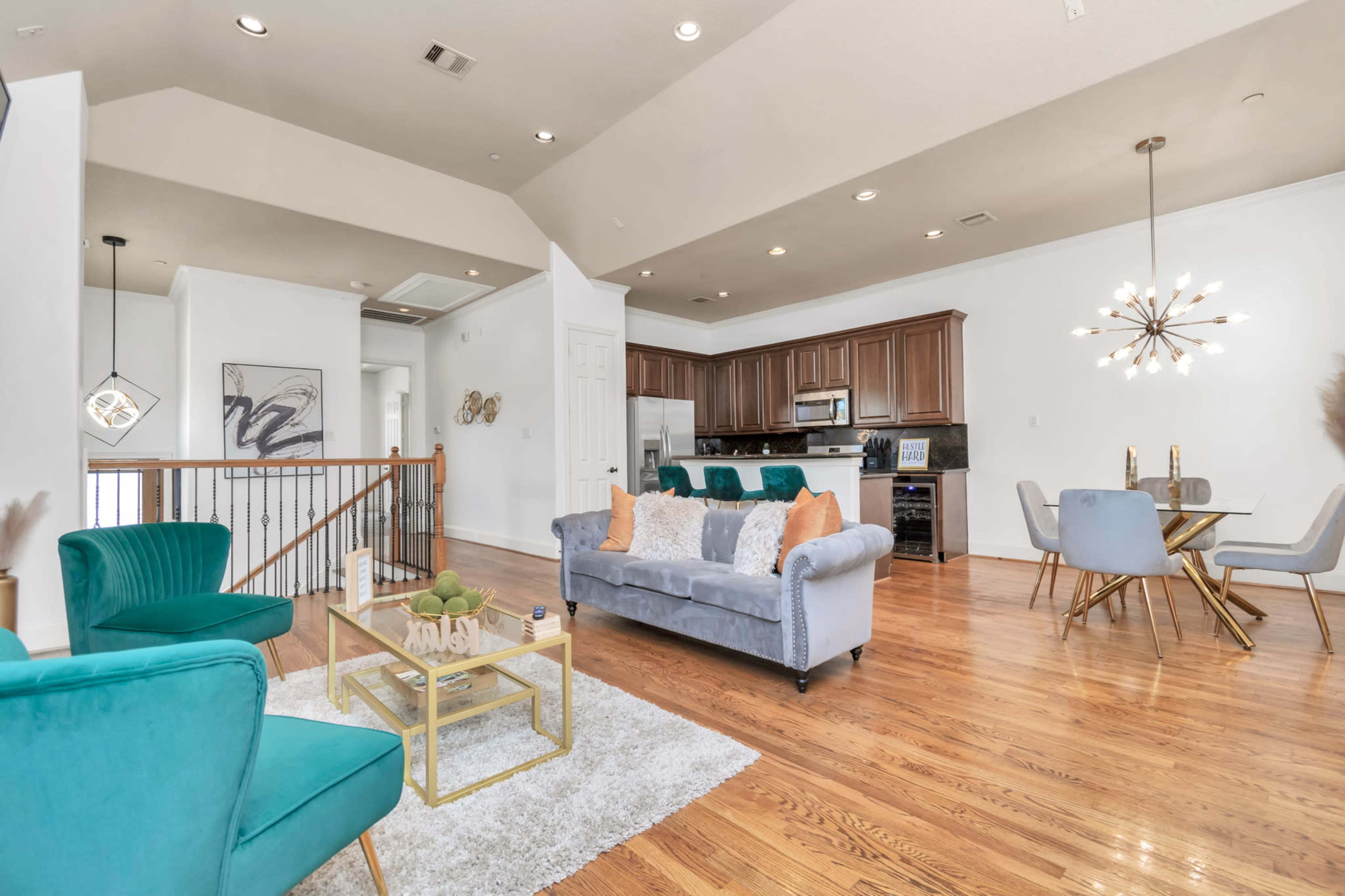 A modern open-concept living area featuring a blue couch, teal accent chairs, and a dining table, with hardwood floors and a kitchen in the background.