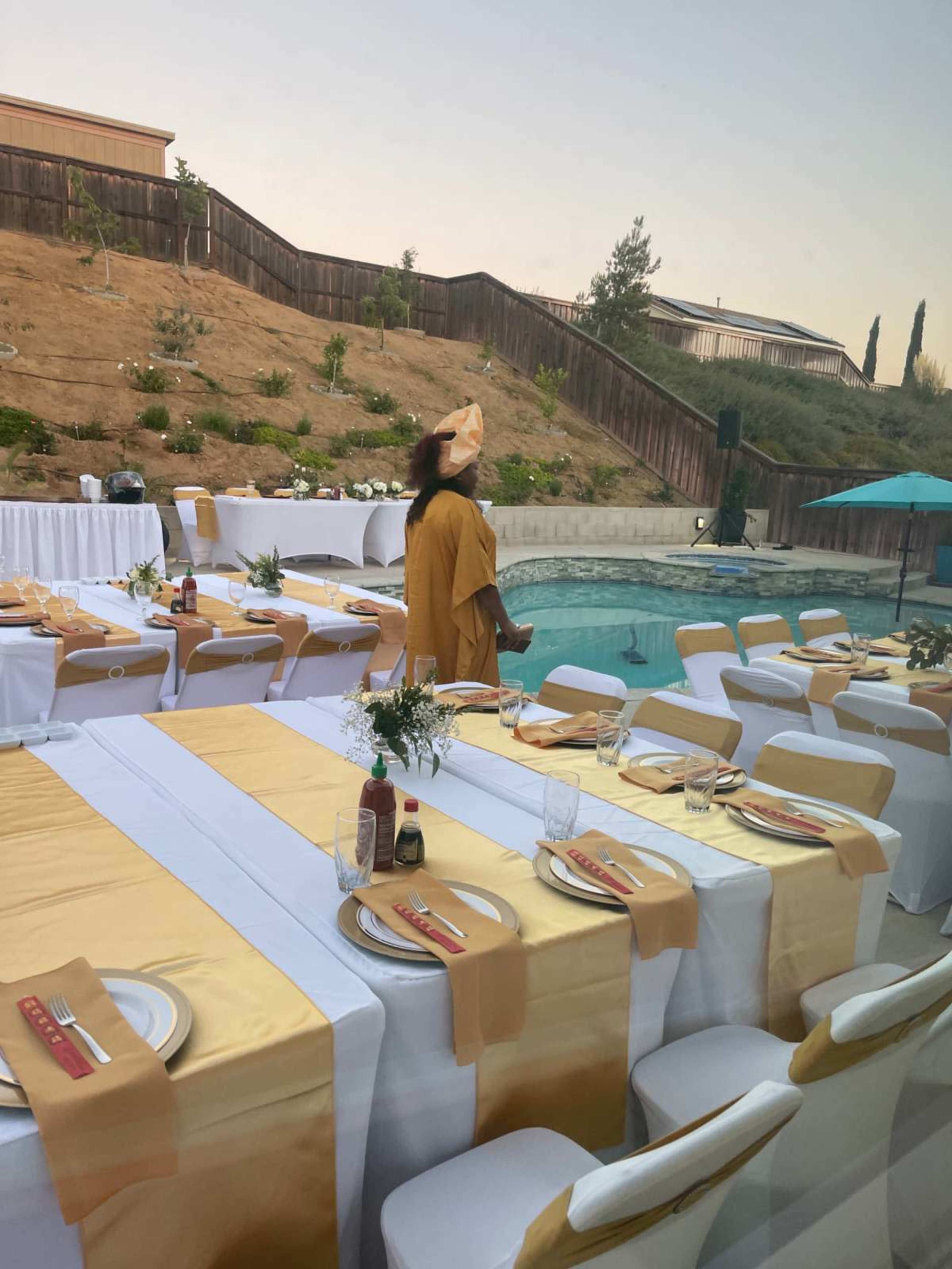 A woman in a yellow outfit stands beside a beautifully set outdoor dining area by a swimming pool, with white and gold table settings.