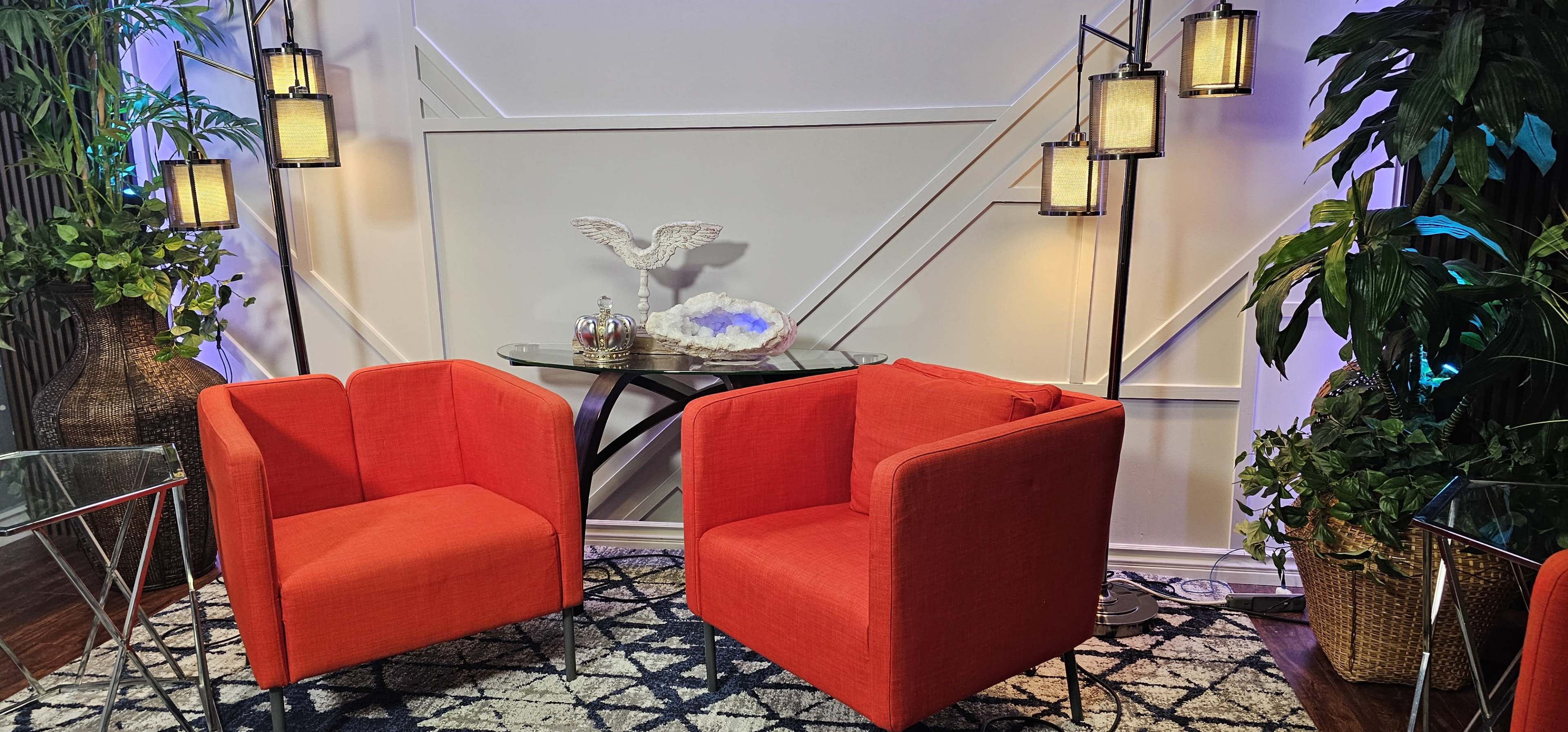 The image shows a cozy seating area with two red chairs, a glass table, and decorative plants in an interior space.