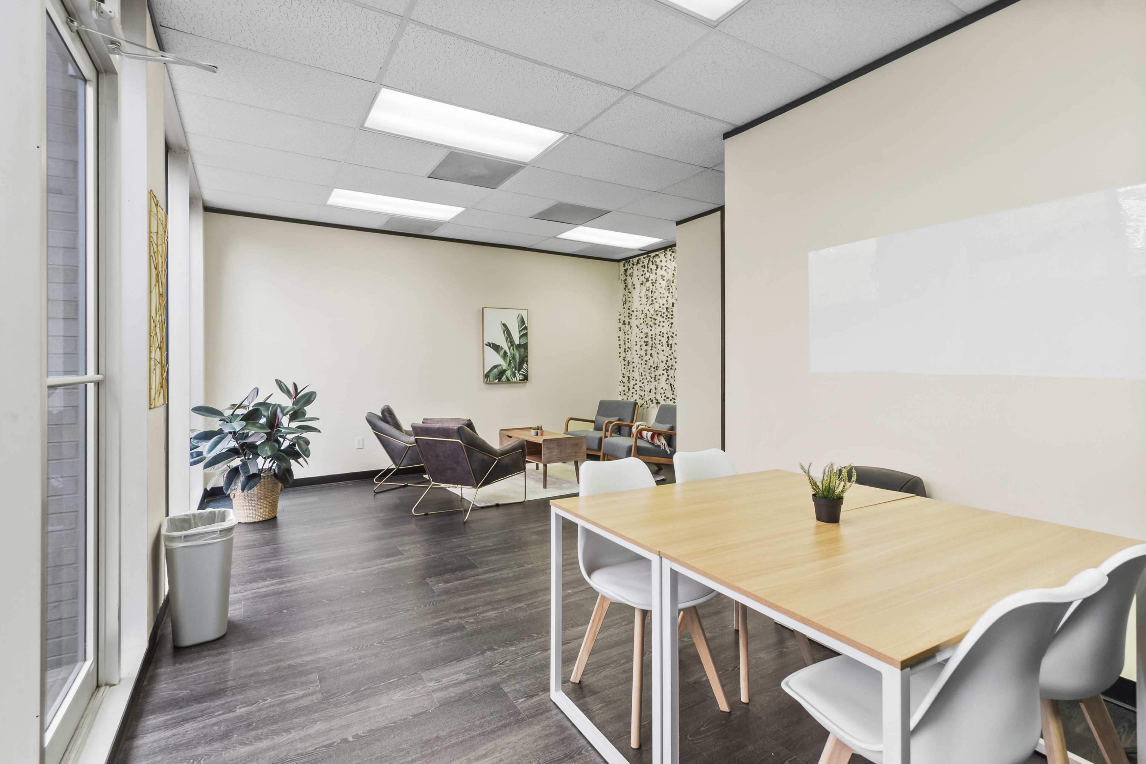 A furnished office space features a wooden table with chairs, a seating area with couches, and greenery against neutral-colored walls.