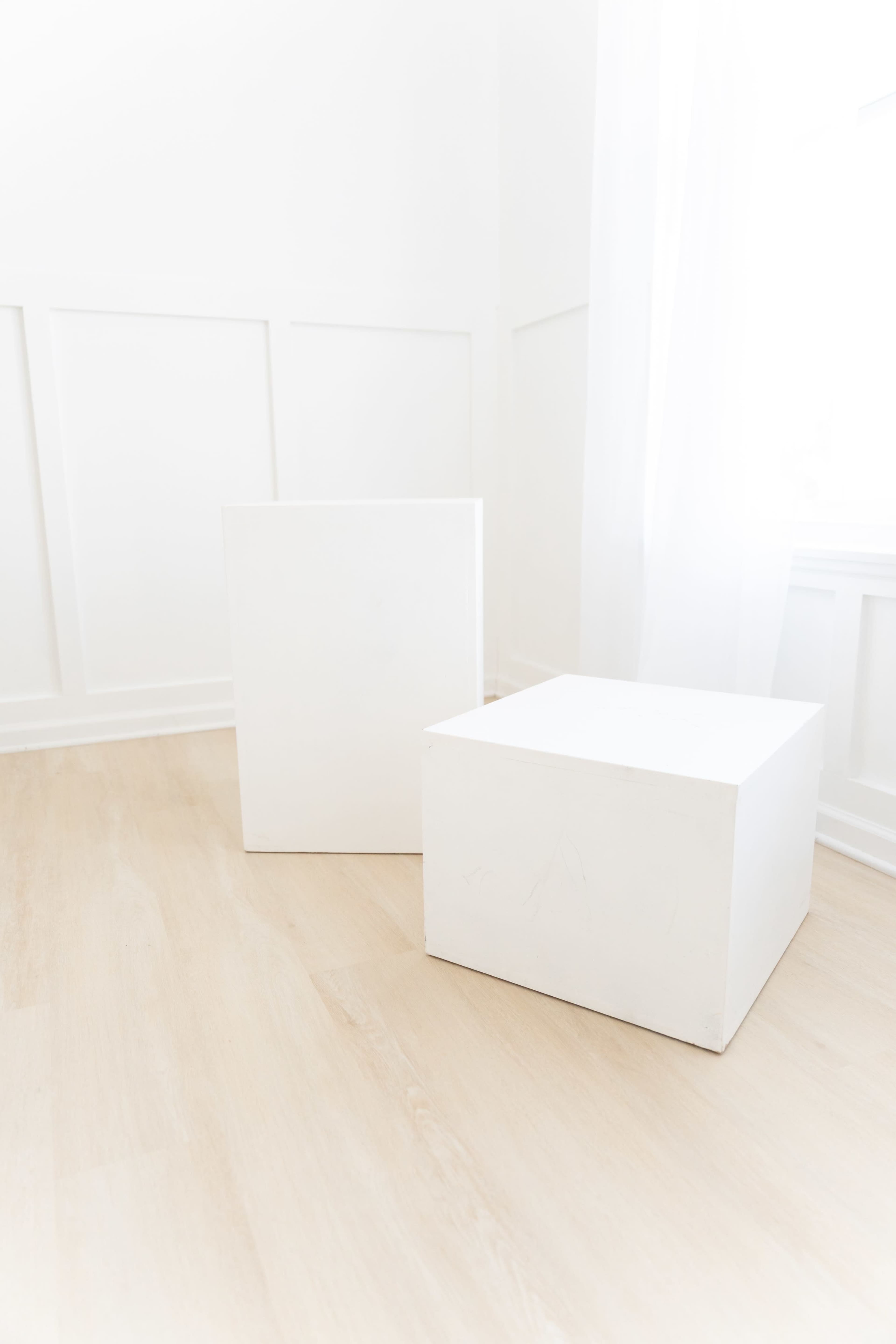 The image shows two white geometric boxes placed on a light wooden floor in a bright, minimalist room.