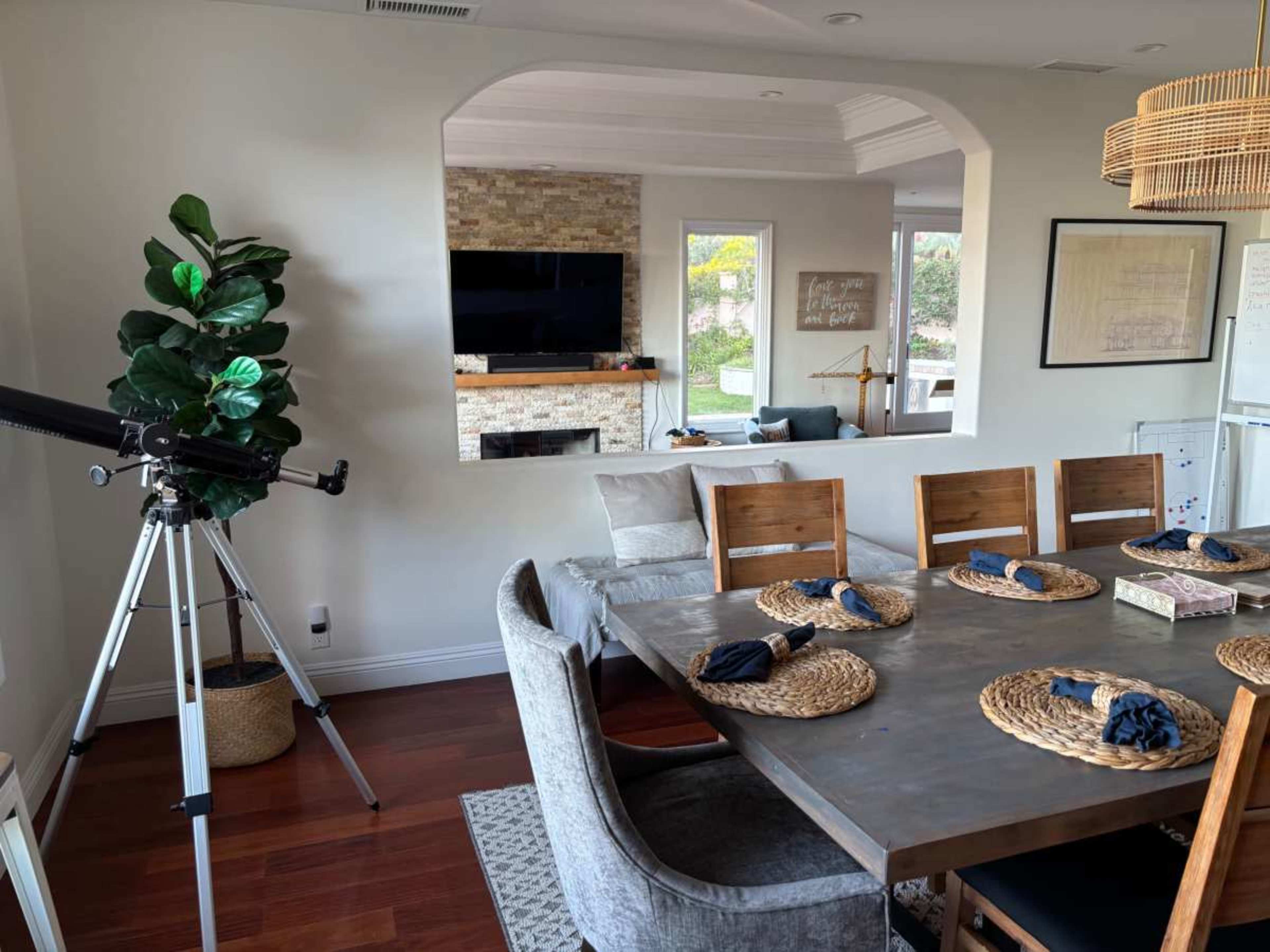 The image shows a dining area with a large table set with decorative placemats and utensils, a telescope positioned nearby, and a view of a living space with a television and a fireplace in the background.