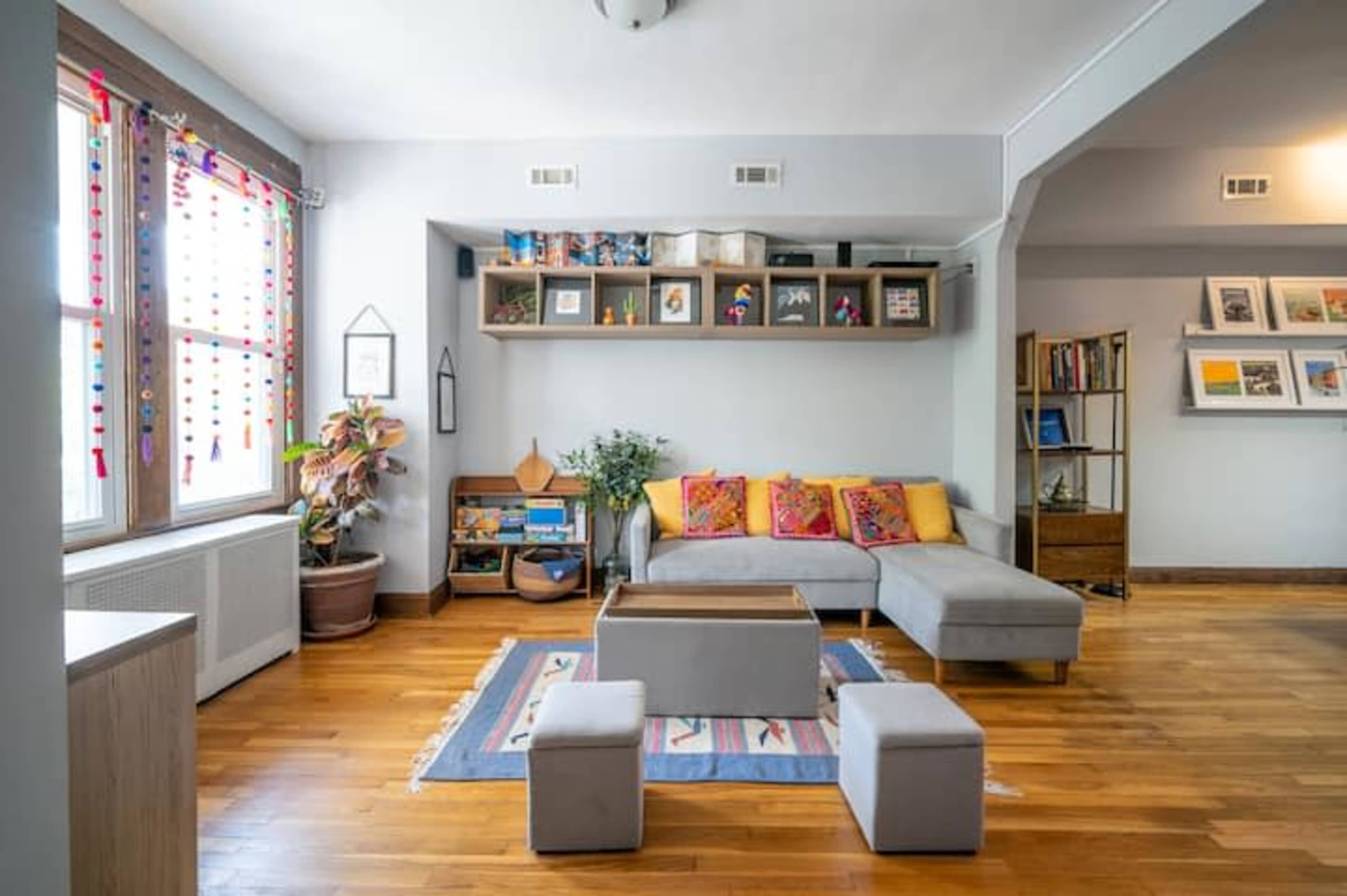 The living room features a gray sectional sofa with colorful pillows, a wooden coffee table, and a bookshelf against the wall, with two small stools on a patterned rug.
