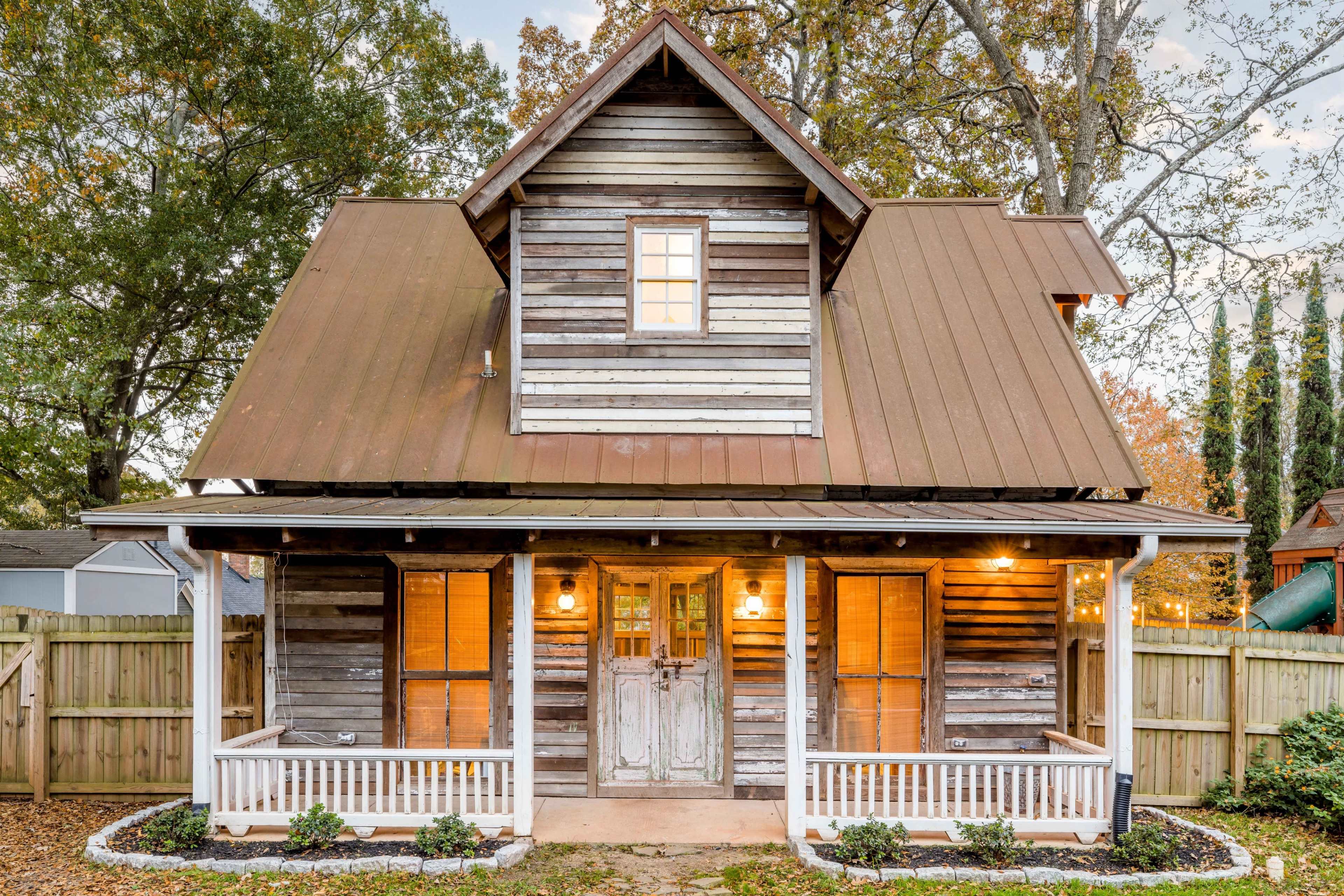A two-story wooden house with a metal roof, featuring a front porch and surrounded by a wooden fence and greenery.