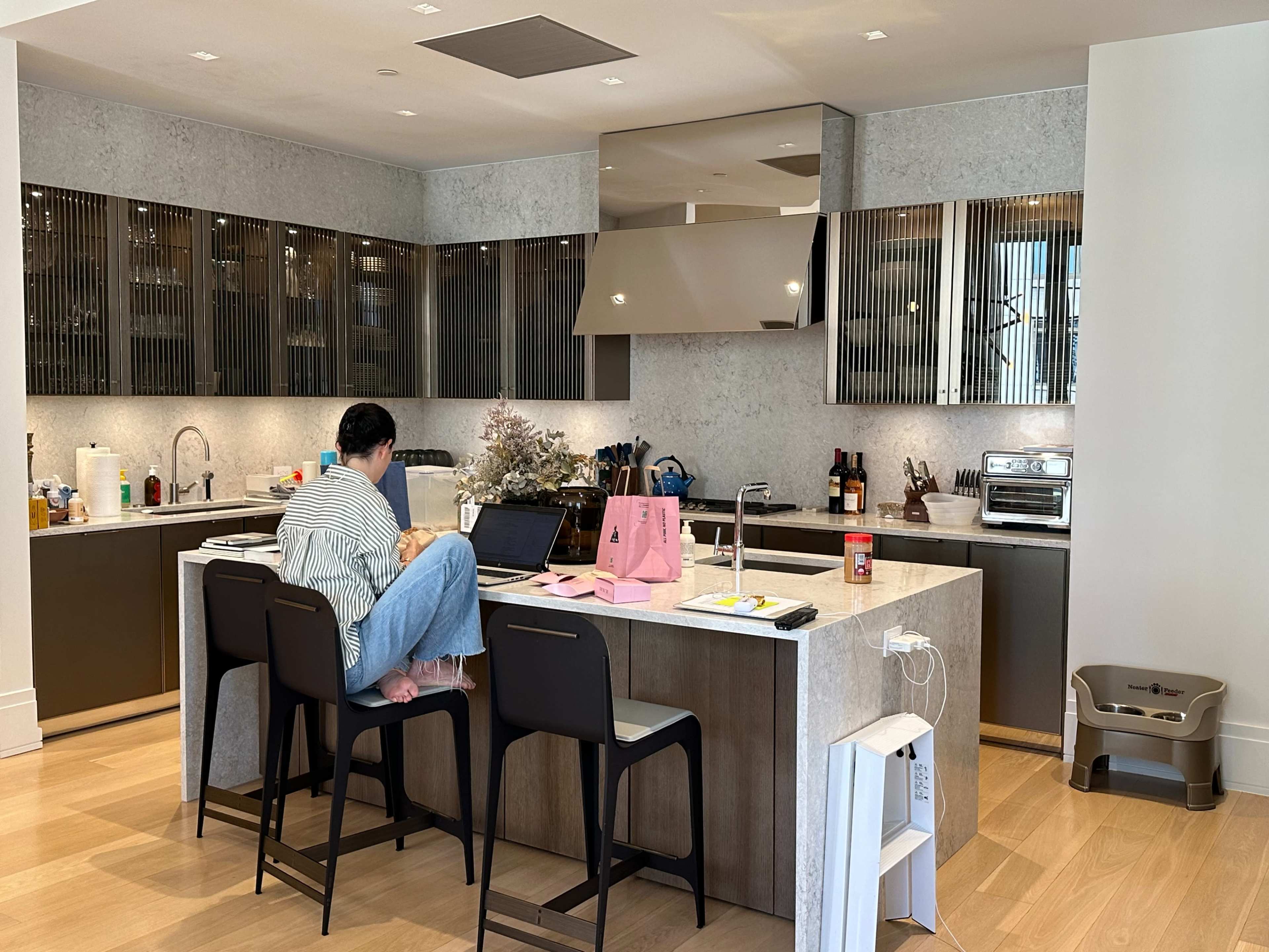 A person works on a laptop while sitting at a kitchen island with modern cabinetry and appliances.