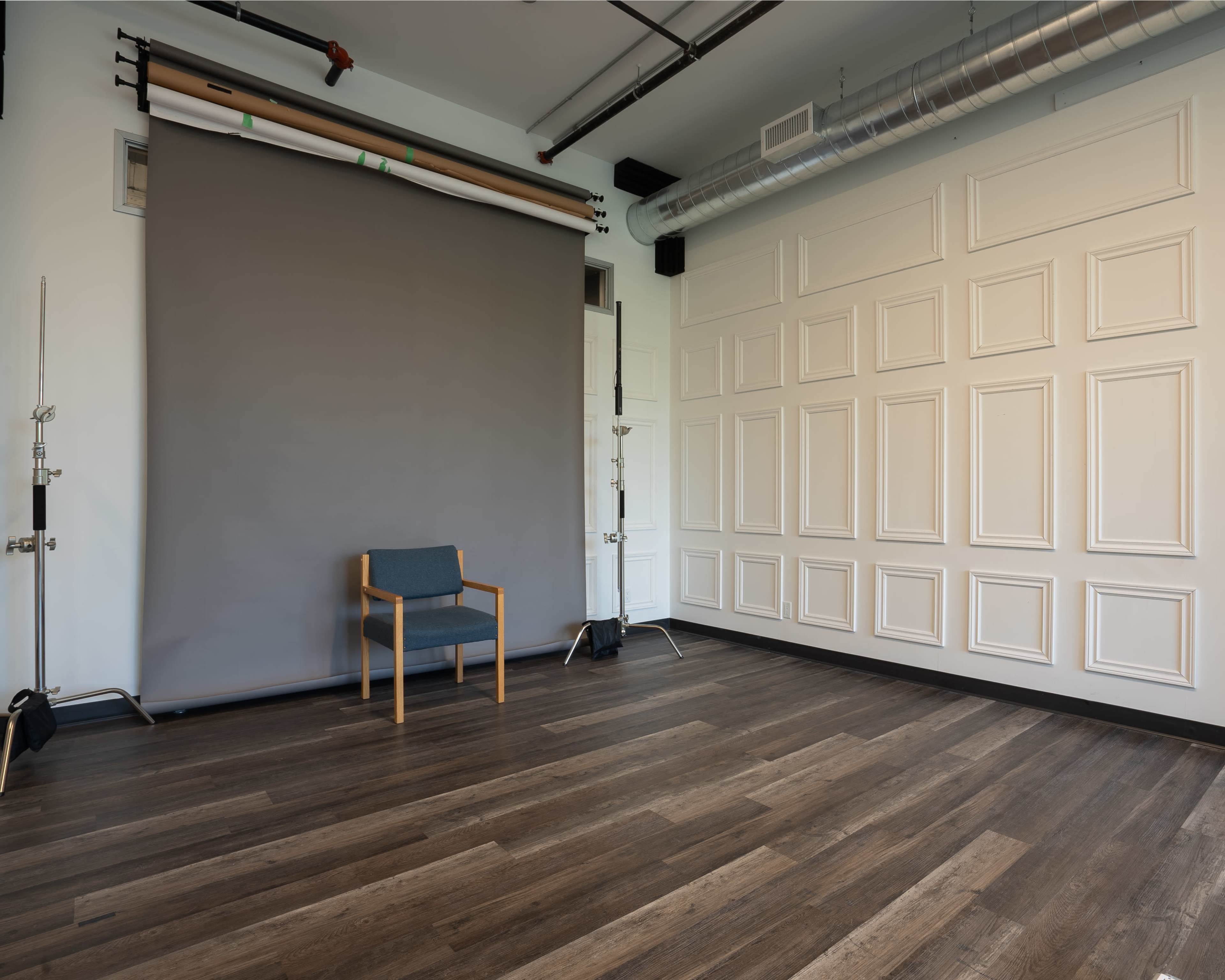 A photo studio with a gray backdrop, a wooden floor, and a single chair positioned in front of the backdrop.