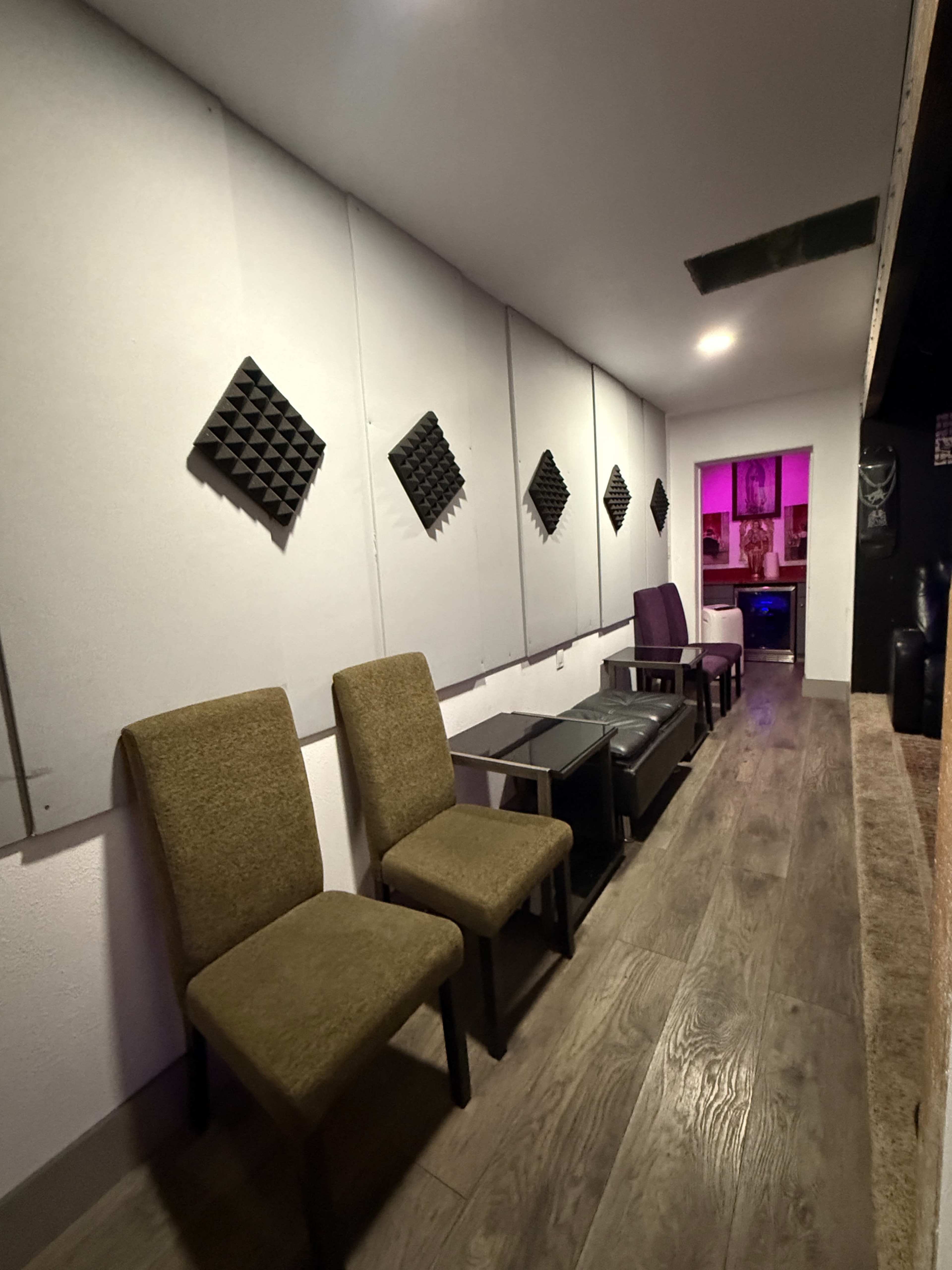 The image shows a narrow room with soundproofing panels on the walls, featuring two dark chairs lined up against one side and a purple chair at the far end.