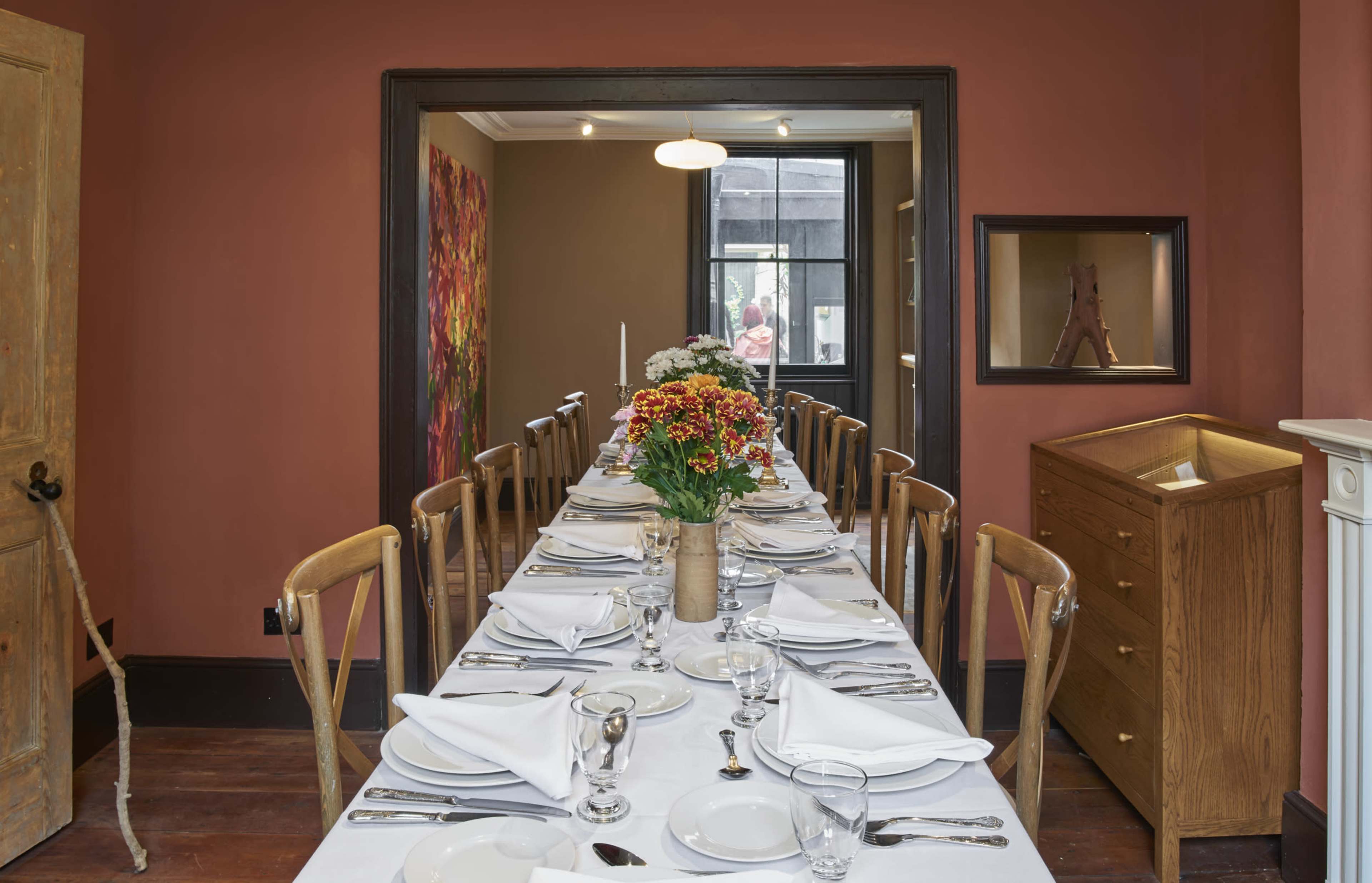 A long dining table is set with white linens, utensils, and a floral centerpiece in a warmly colored room with decorative art on the walls.