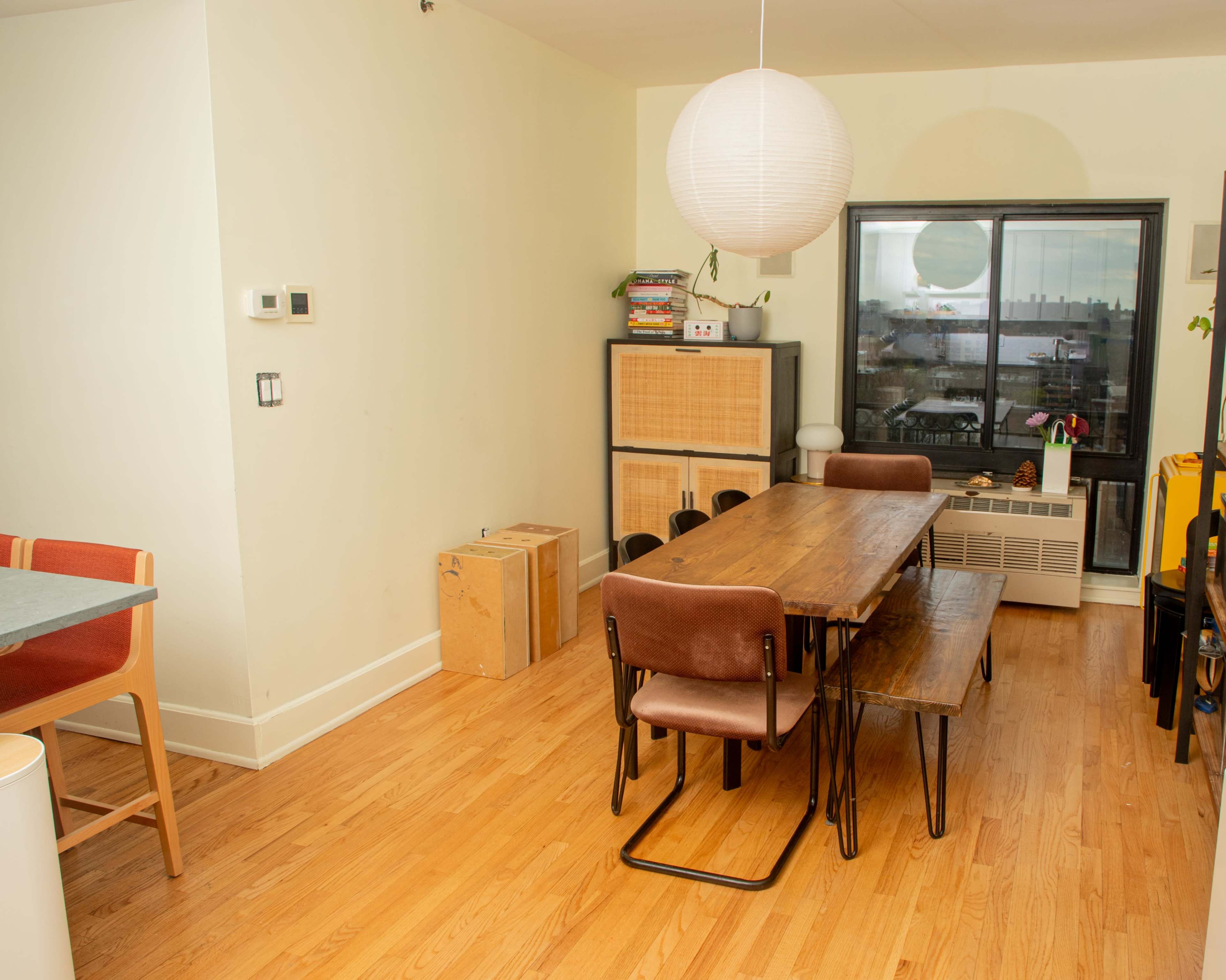 The image features a dining area with a wooden table surrounded by chairs, a large window, and a minimalist decor.