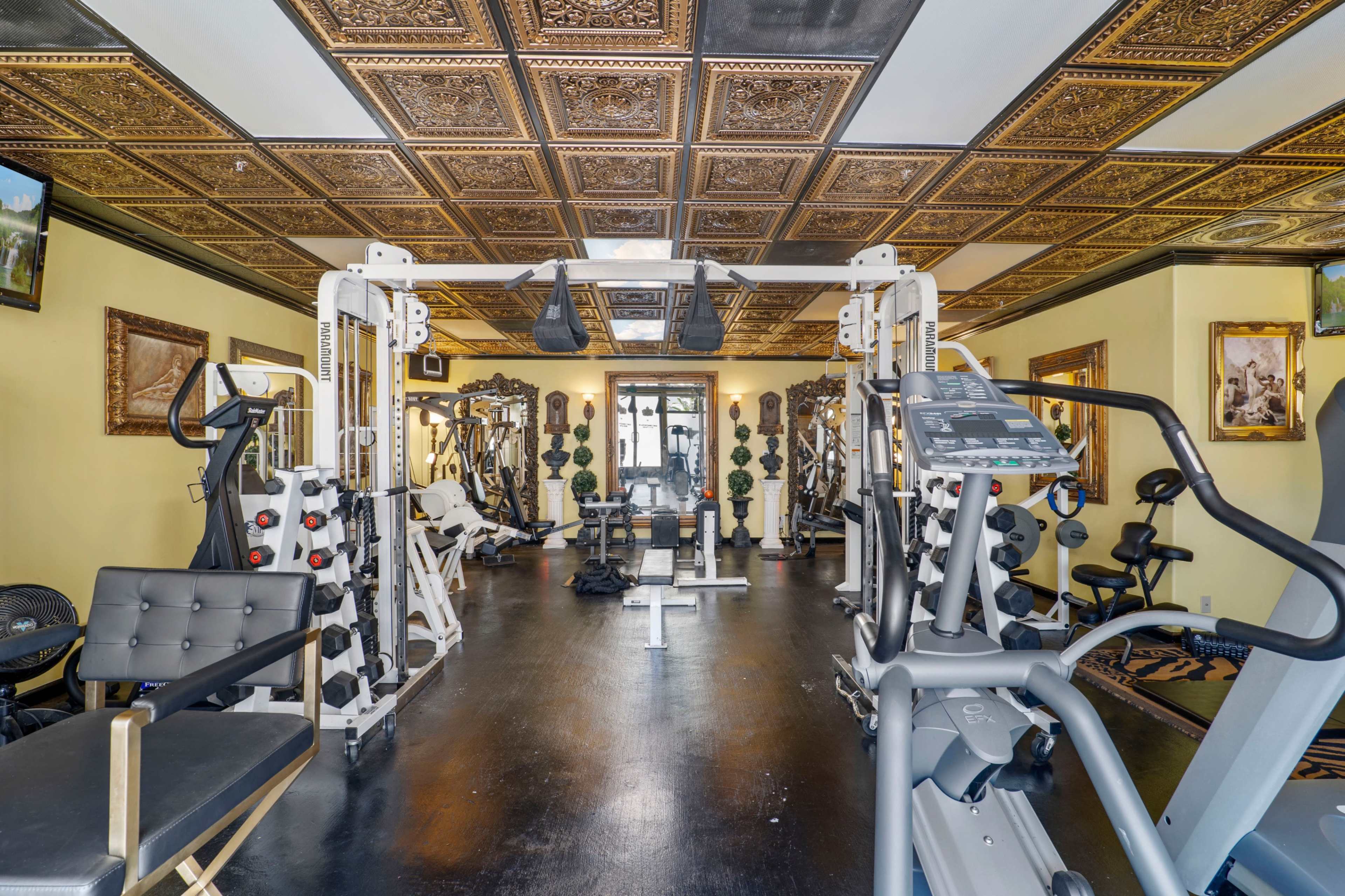 A gym with various exercise machines, illuminated by wall-mounted artwork and decorative ceiling tiles.