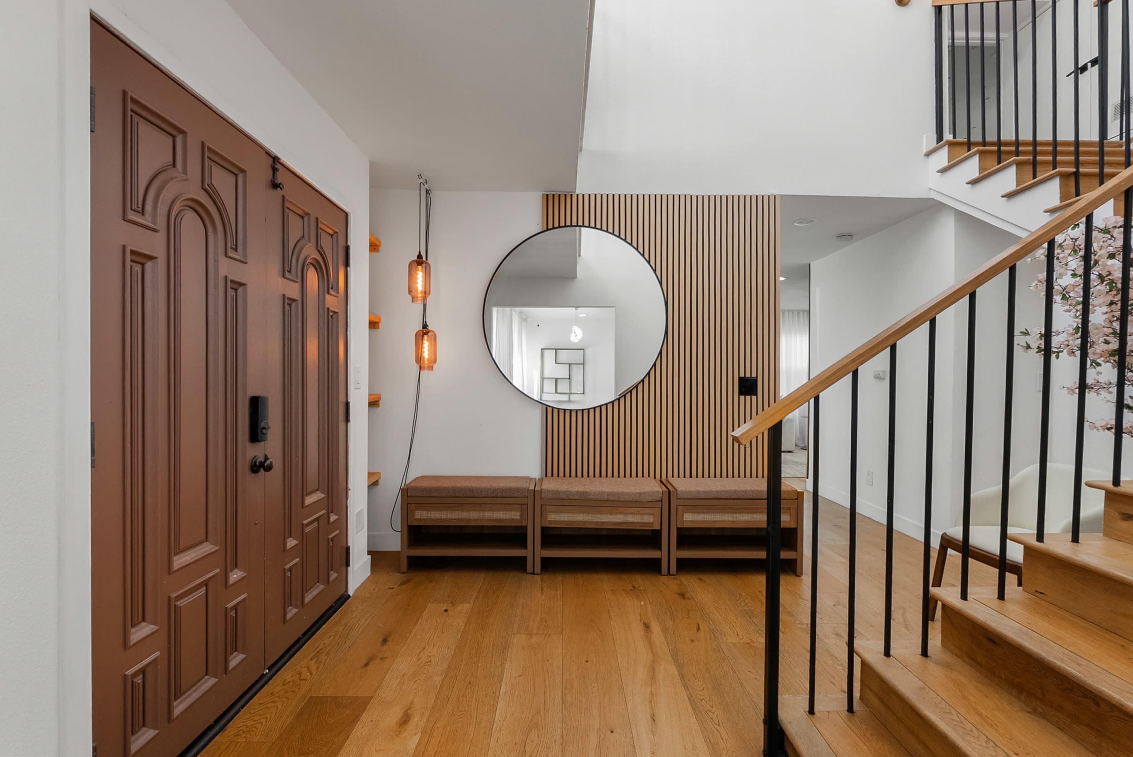 The image depicts an entrance hallway featuring a pair of brown doors, a round mirror, wooden benches, and a staircase with a wooden railing.
