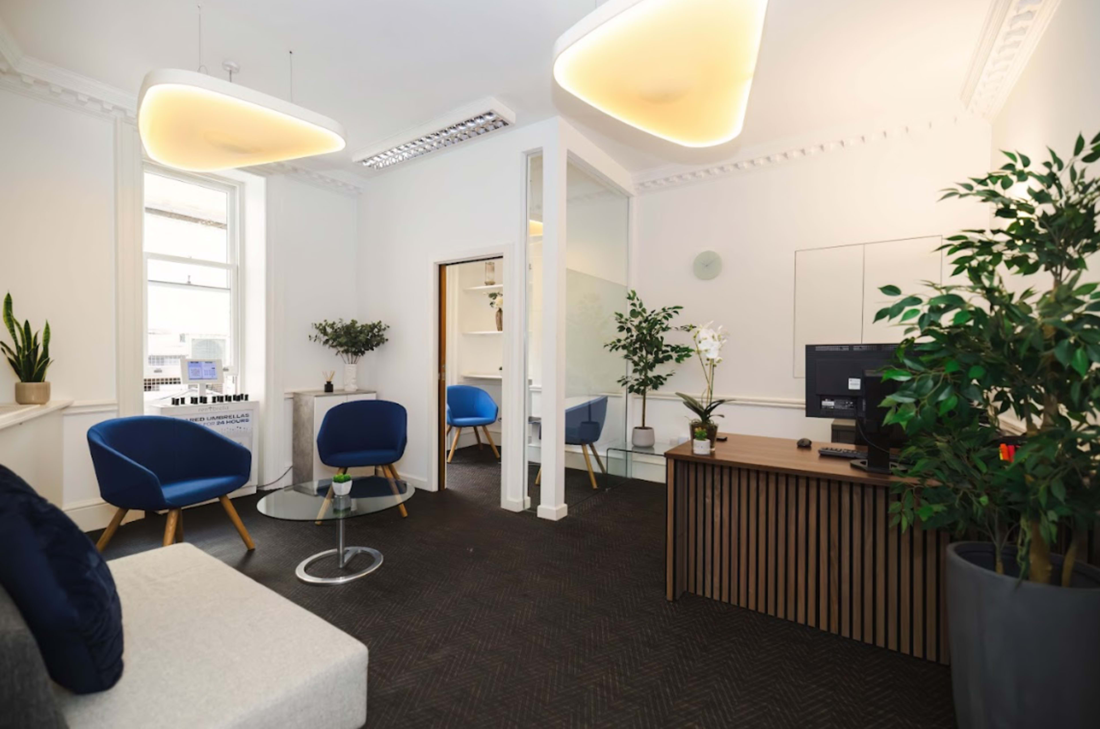 The image shows a modern office space with two blue chairs, a glass coffee table, a couch, a desk with a computer, and plants in each corner.