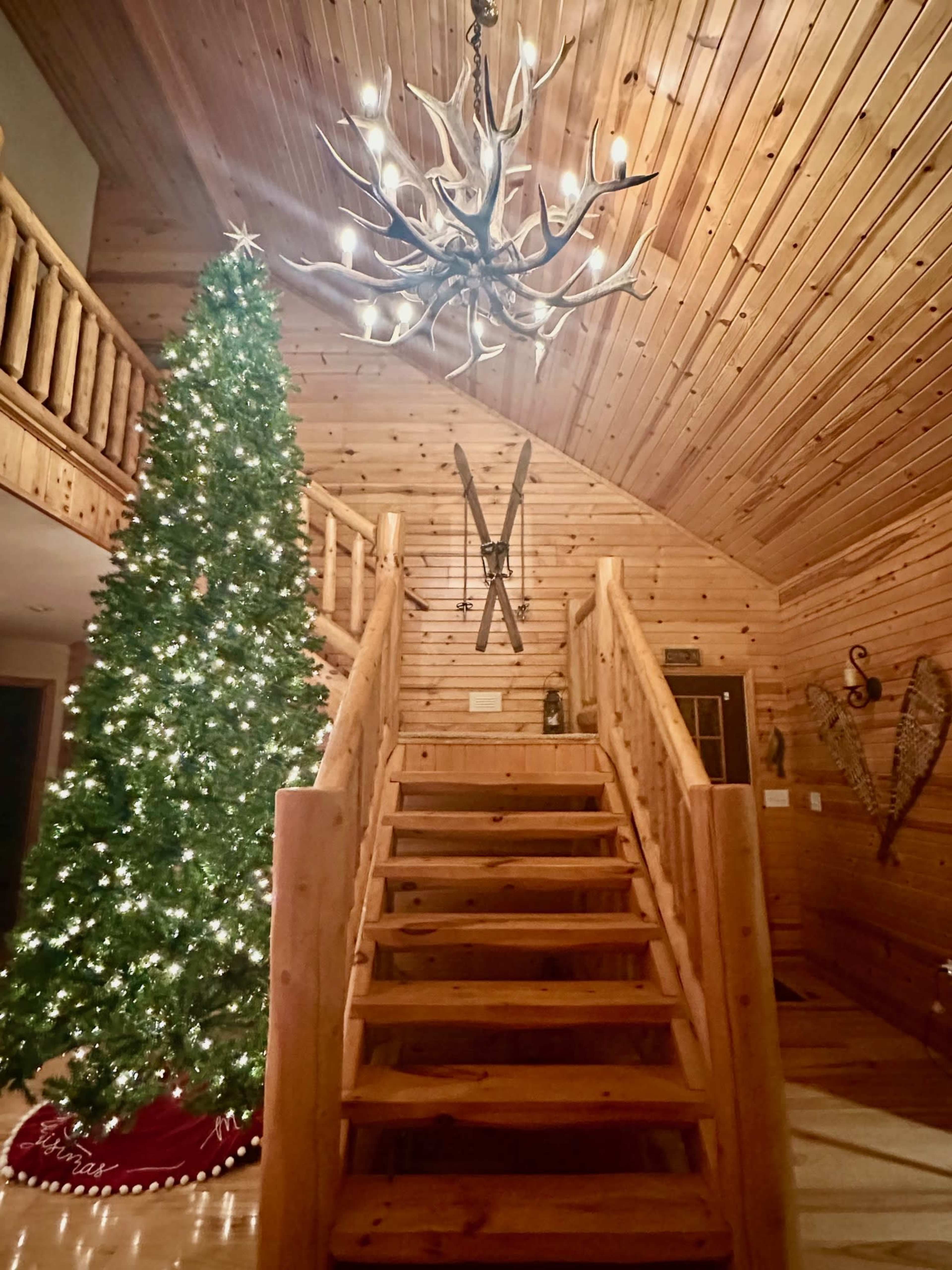The interior of a wooden cabin features a tall Christmas tree with lights near a staircase, a large antler chandelier hanging above, and wooden paneling throughout.