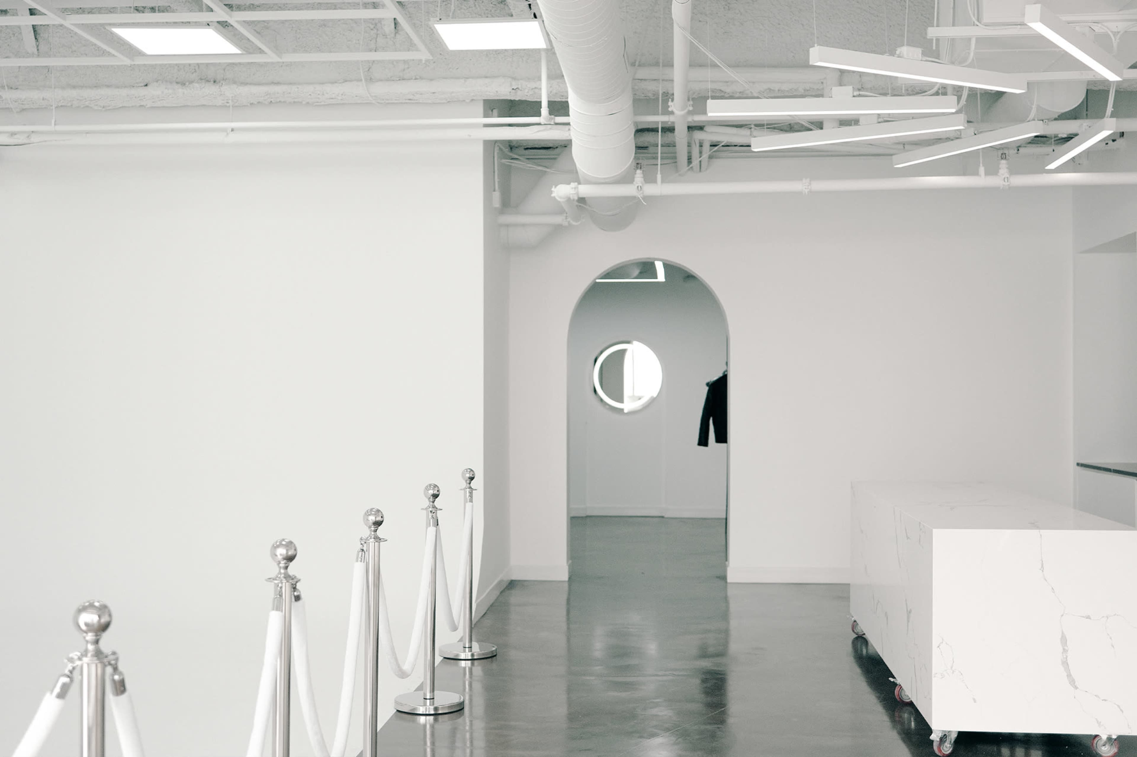 A modern, minimalist interior with white walls, a rounded entrance, a circular window, and a long, rectangular white counter, lined by stanchions and ropes.