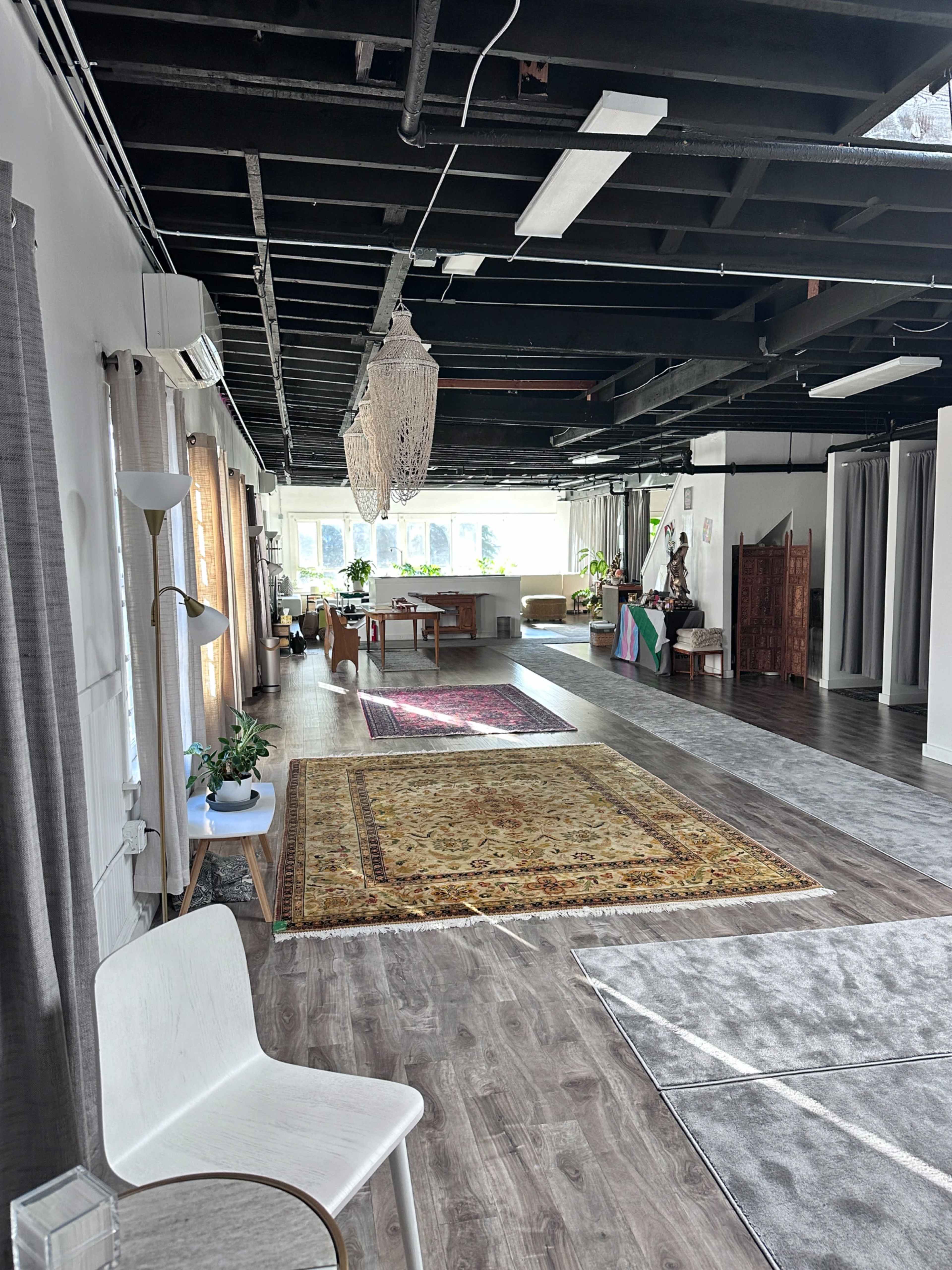 The image shows a spacious, well-lit interior with hardwood flooring, decorative rugs, plants, and various furniture pieces, including a white chair and a table.