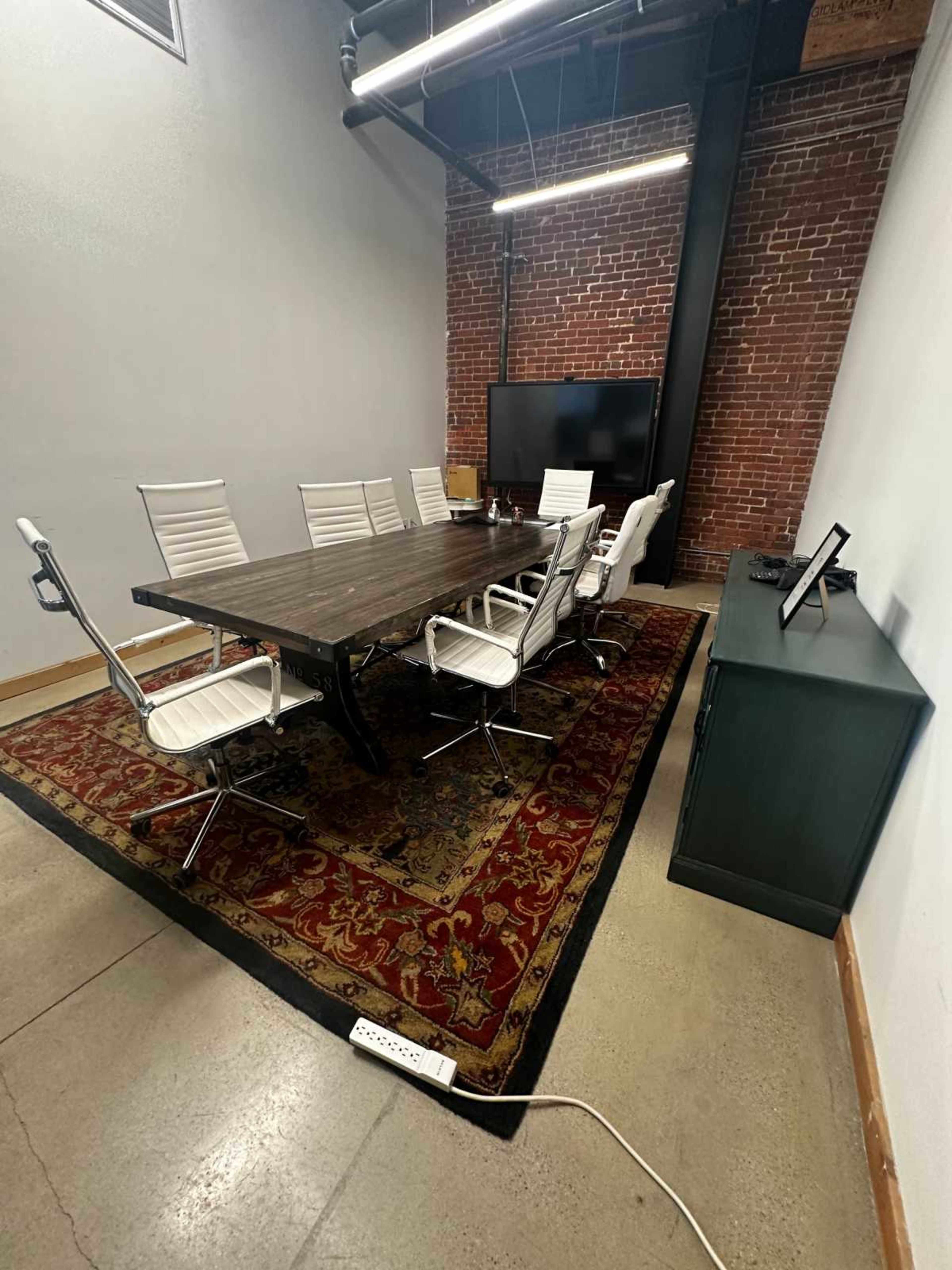 The image shows a conference room with a large wooden table surrounded by white chairs on a patterned rug, and a television mounted on a brick wall.
