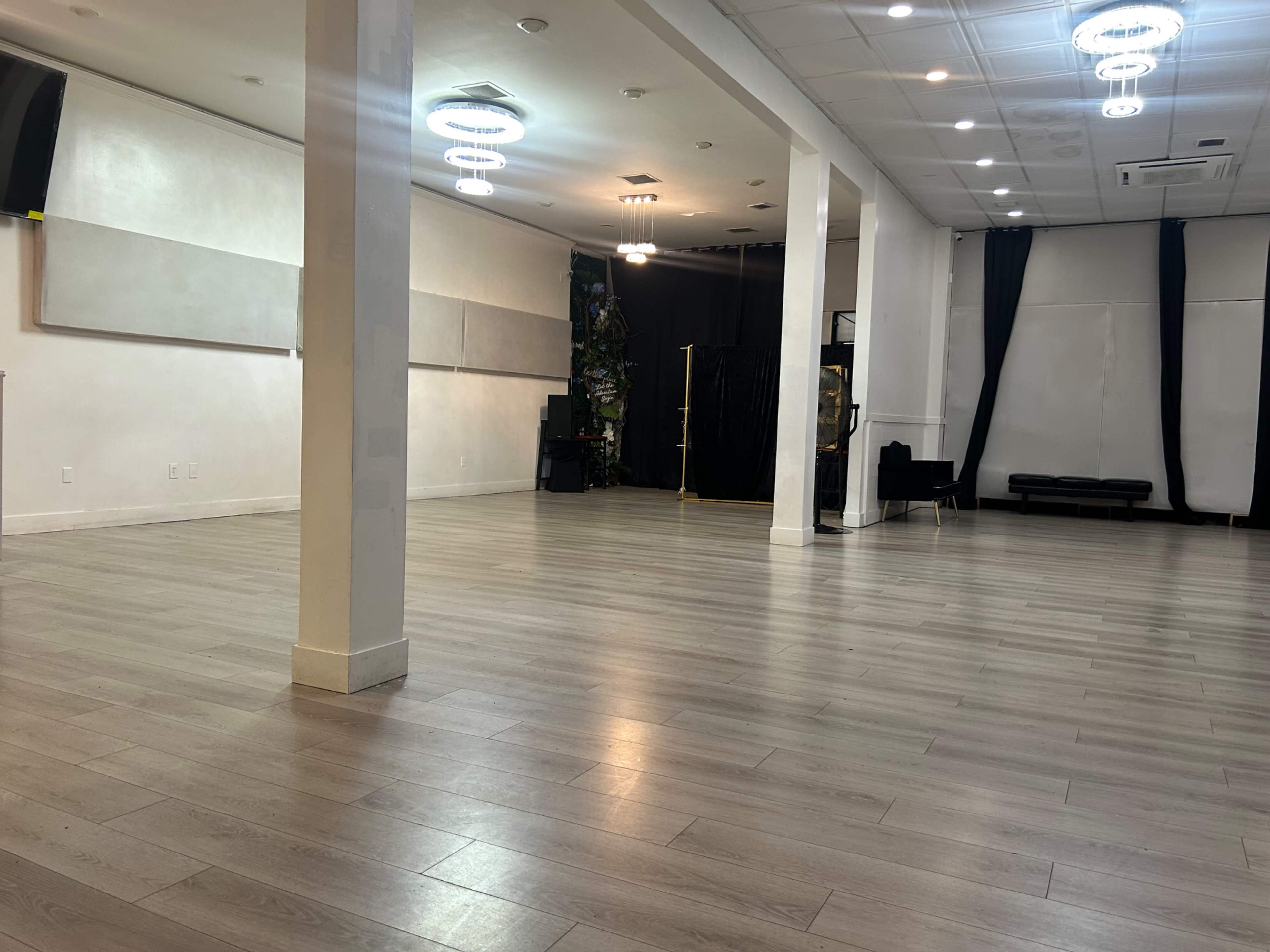 The image shows a spacious, empty room with wooden flooring, white walls, and modern light fixtures hanging from the ceiling.