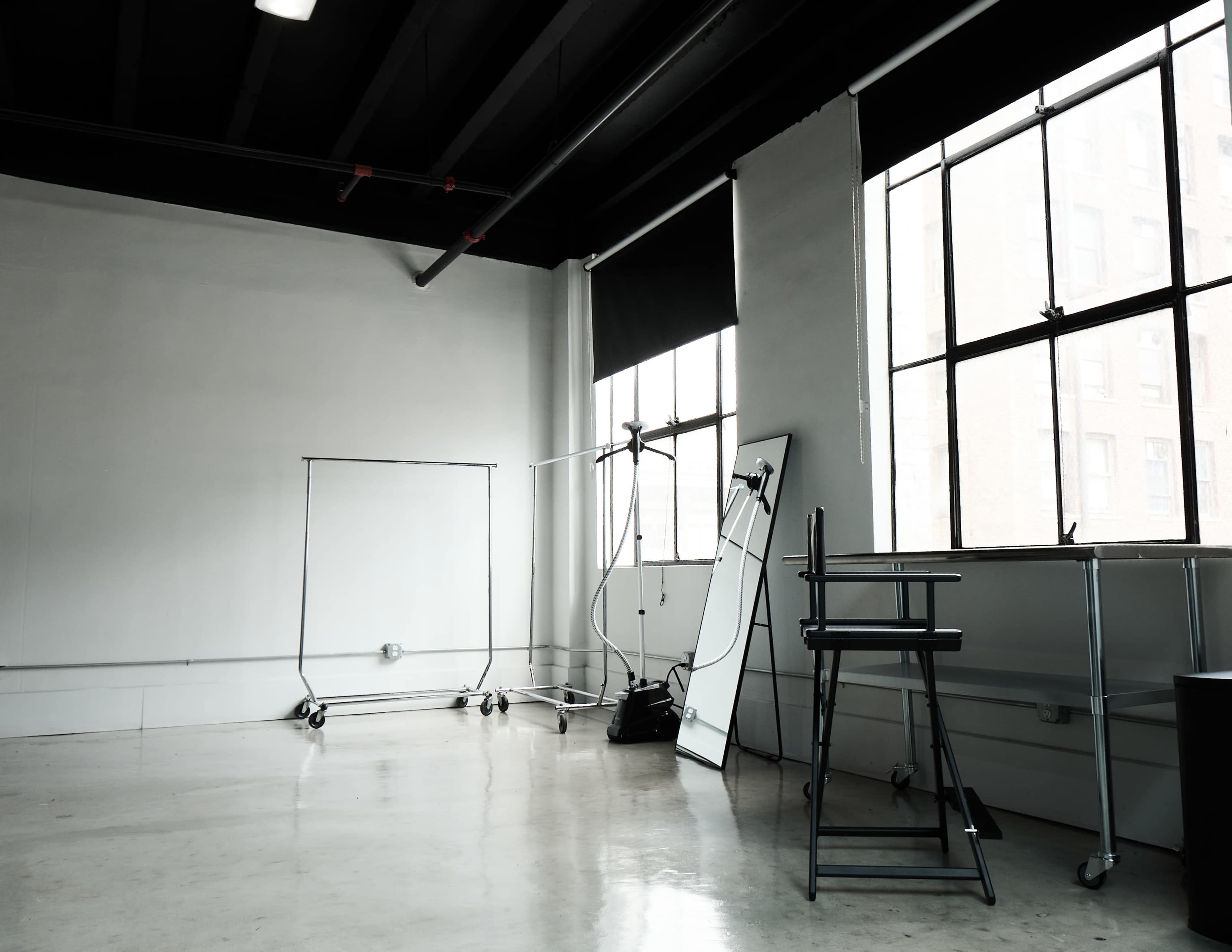 An empty studio space features large windows, a mirror, clothing racks, and a stool against a concrete floor.