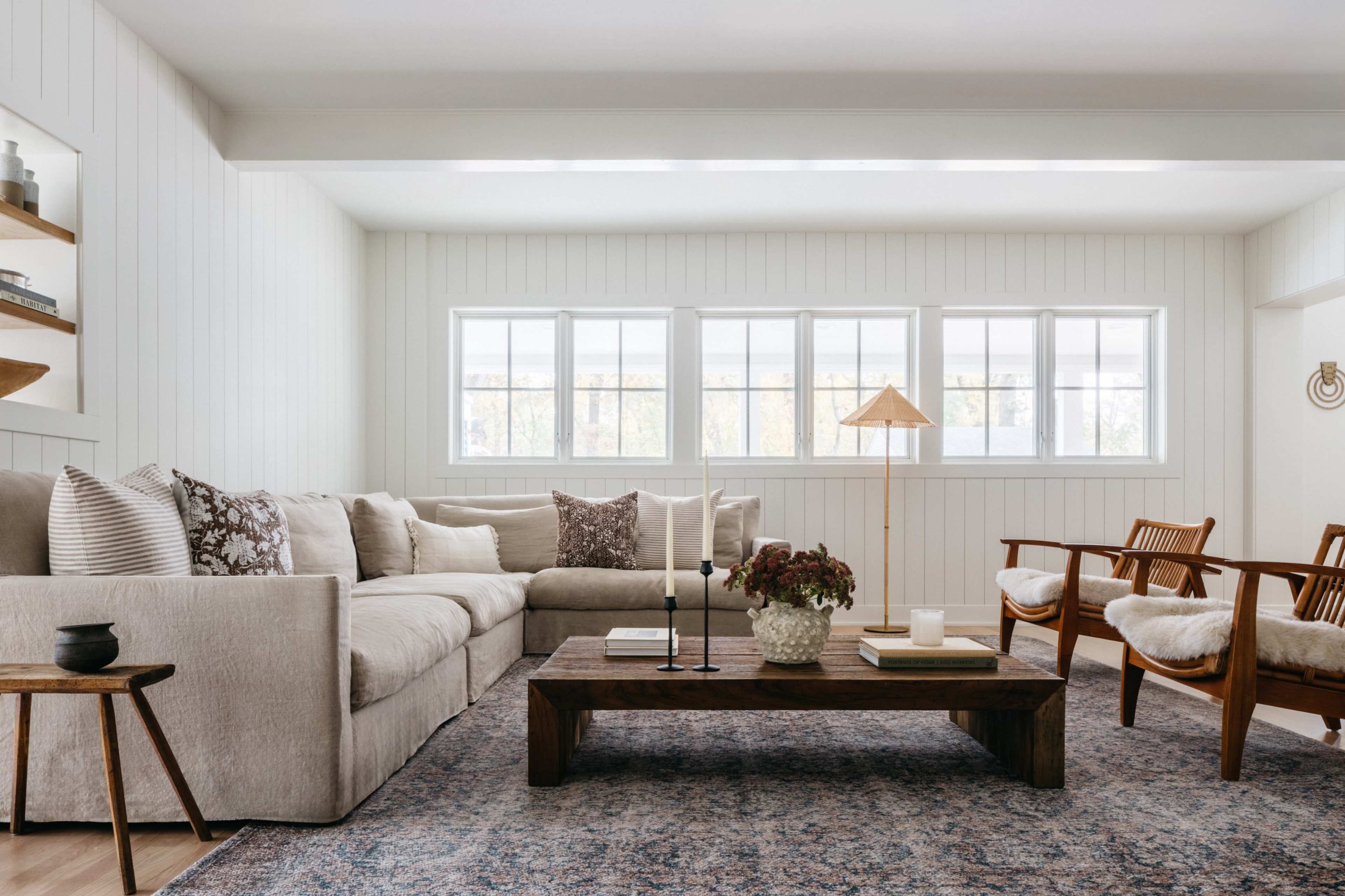 A cozy living room with a large beige sectional sofa, a wooden coffee table, and two armchairs, illuminated by natural light through multiple windows.